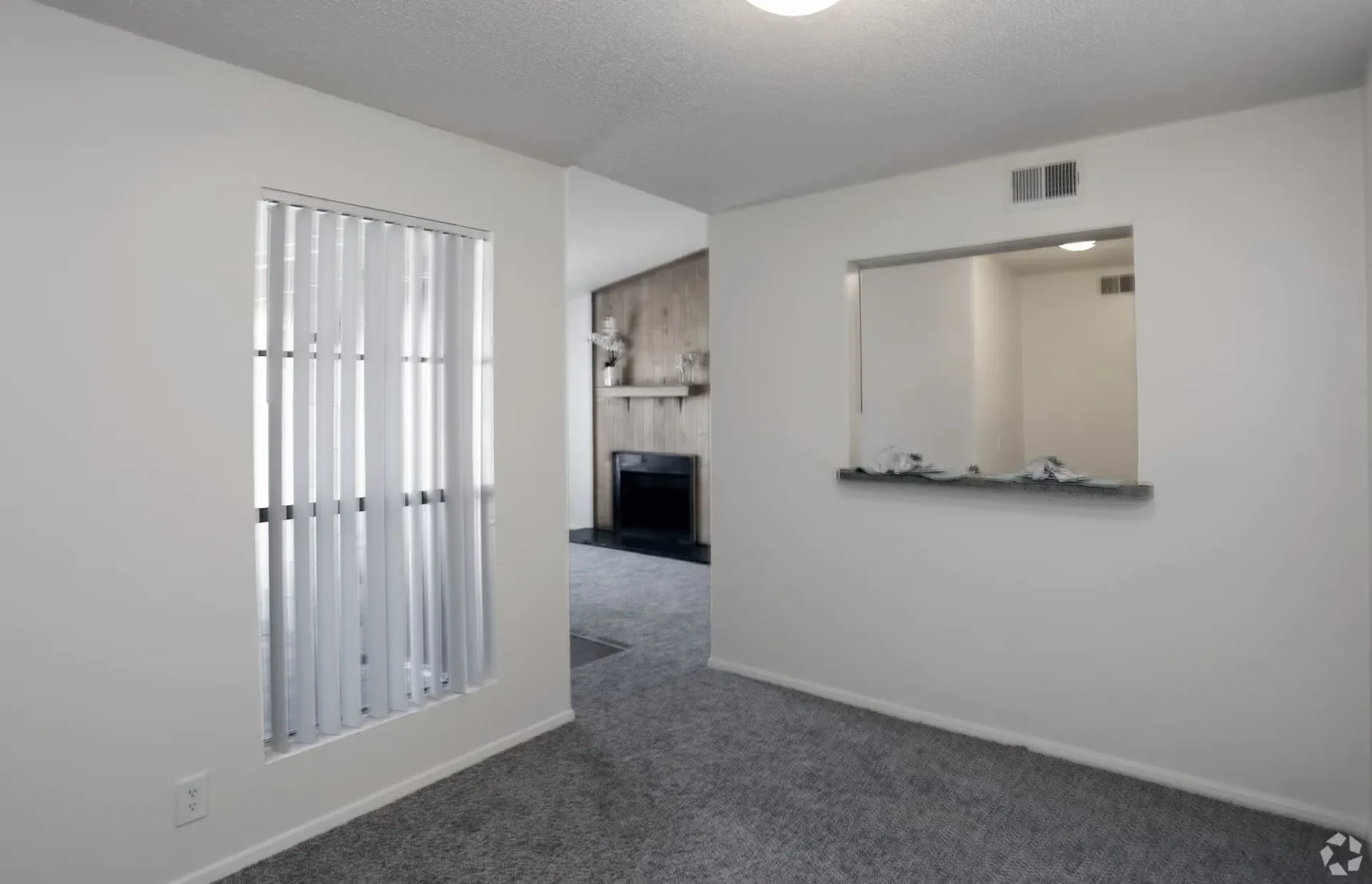 Living room with carpet, vertical blinds, and a pass-through to the kitchen at Woodview, offers apartments in Odessa, TX.