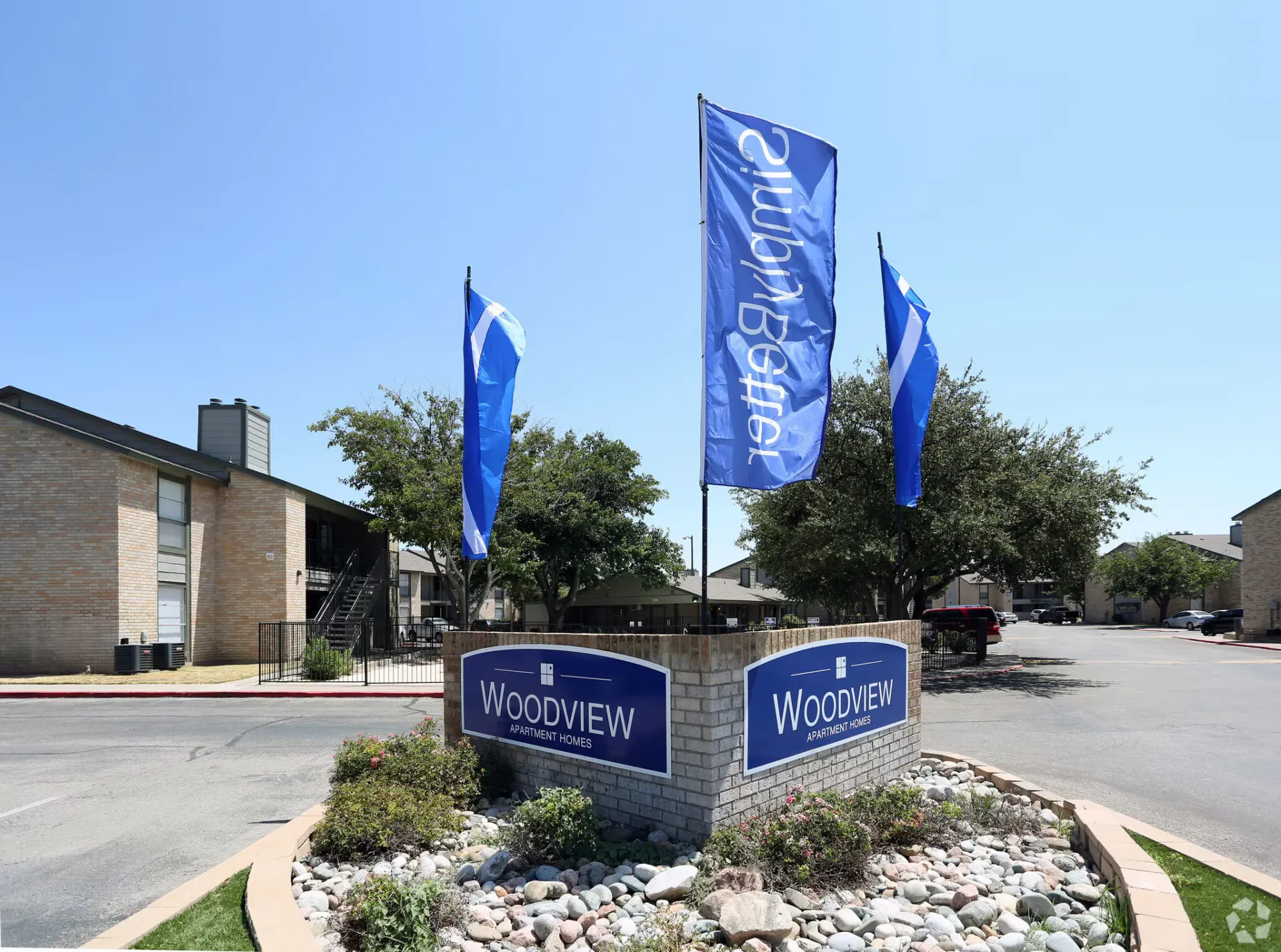 Exterior entrance sign for Woodview Apartment Homes with blue flags and landscaped rock bed.
