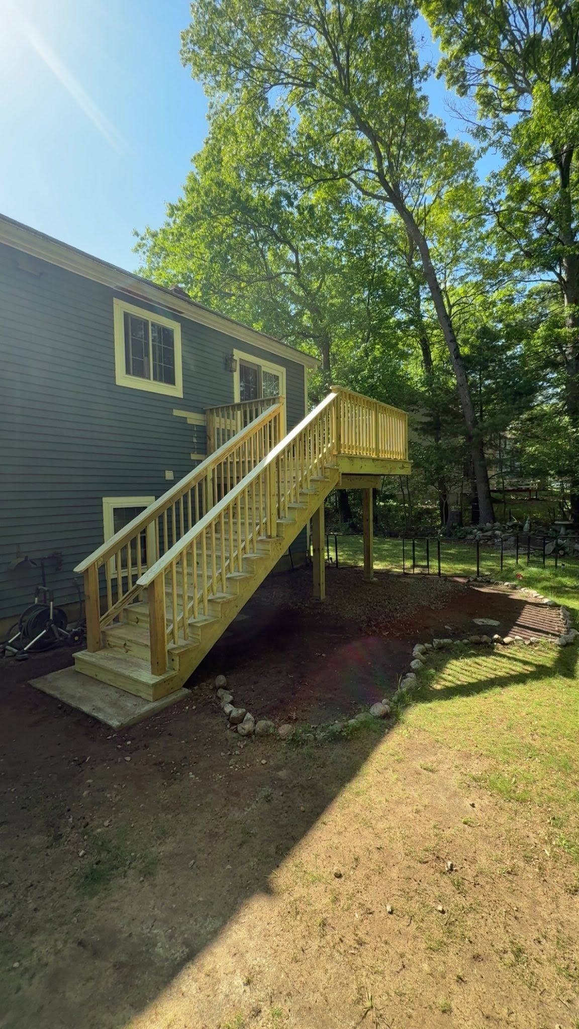 Wooden outdoor staircase with railing leading to a raised deck beside a house, in a sunny wooded yard