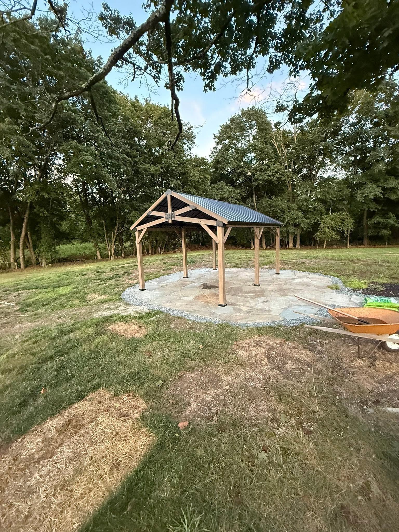 Small open pavilion with a blue roof in a grassy park, surrounded by trees and gravel.