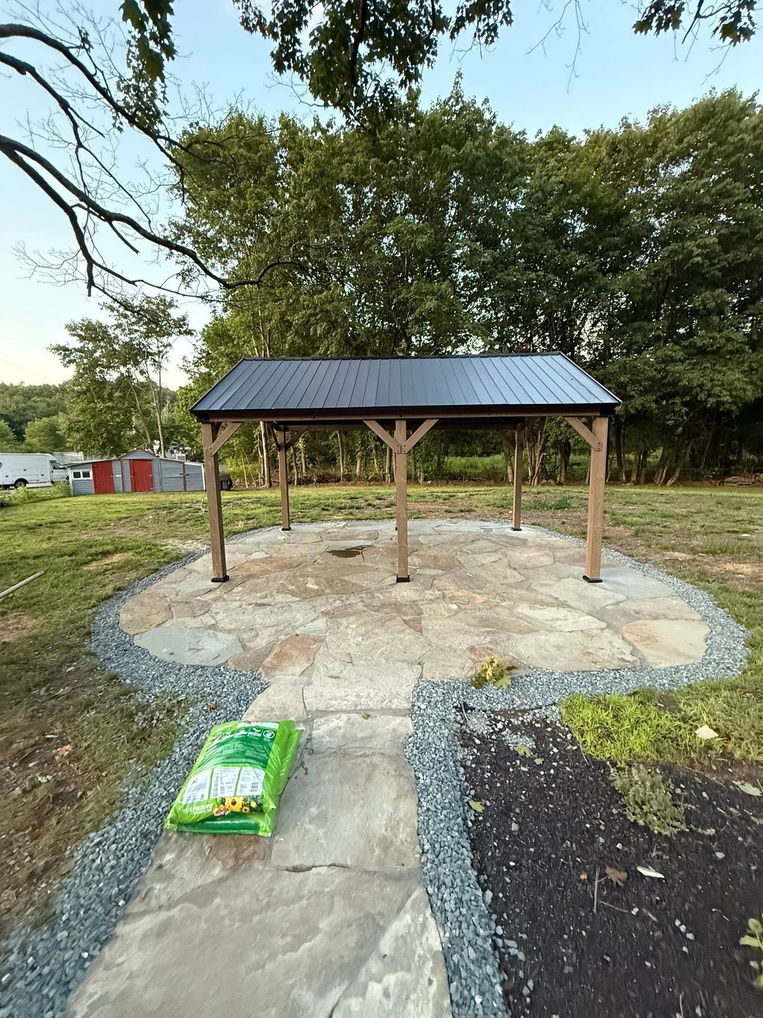 Small wooden pavilion with a metal roof on a gravel pad, beside a concrete path in a grassy park.