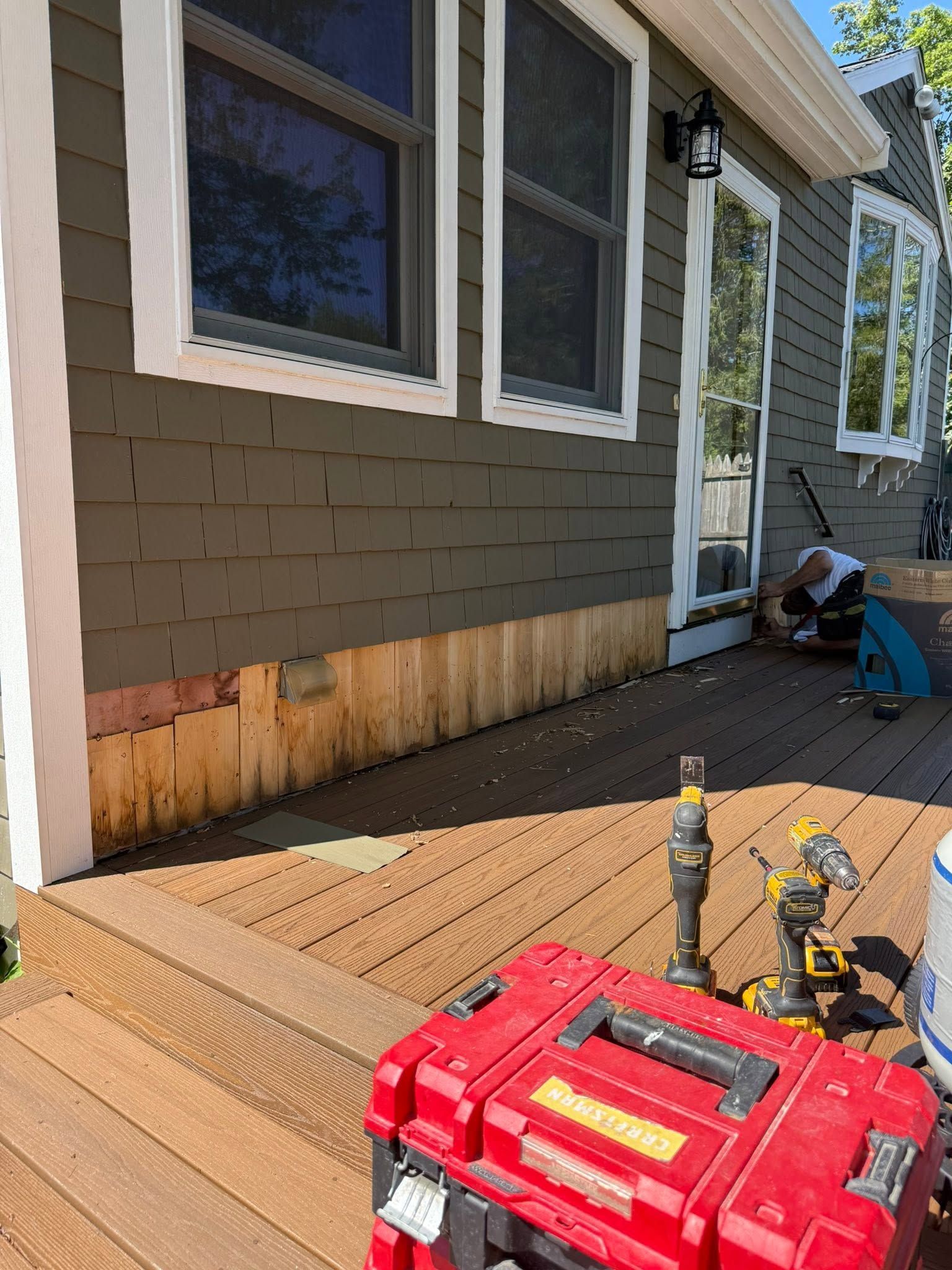 Deck repair tools on a sunlit wooden deck beside a house with gray siding and windows.