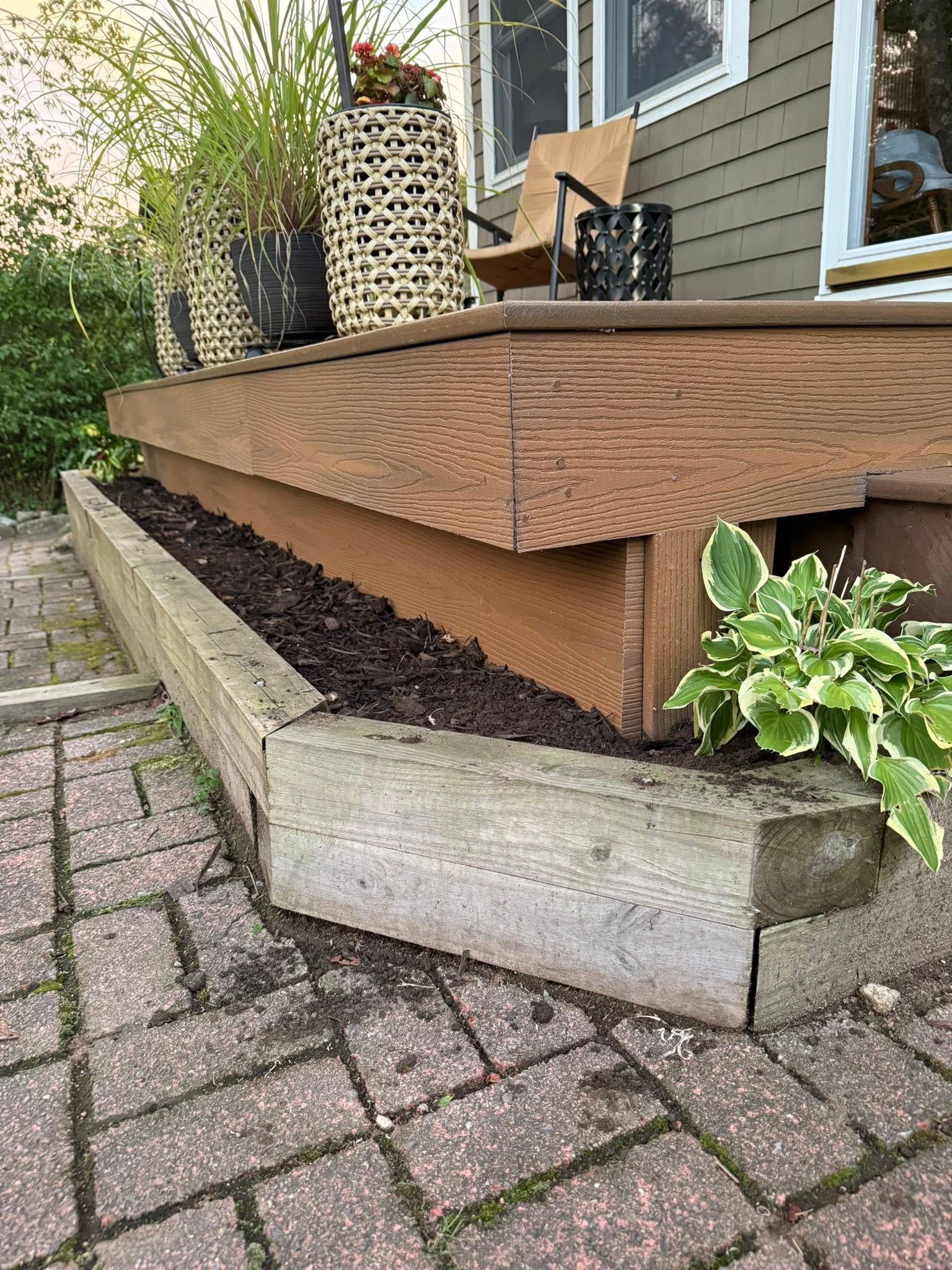 Weathered wooden porch with a concrete planter and green plants beside a brick walkway