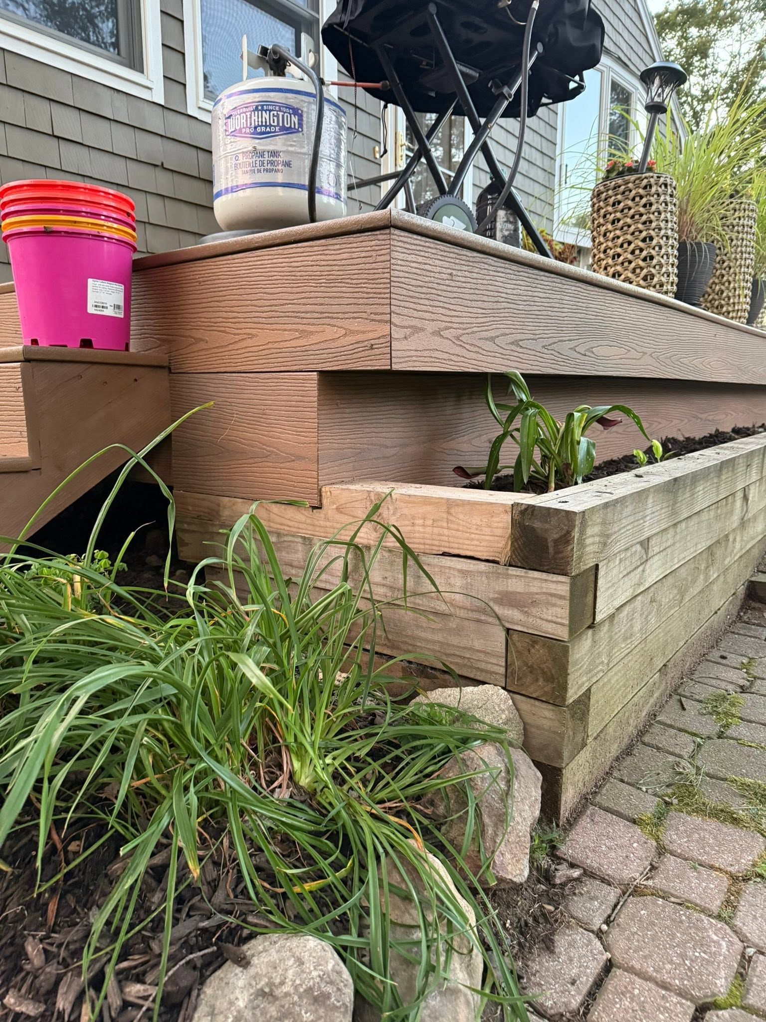 Outdoor wooden planter beside a brick path with green plants and pink buckets on the deck steps