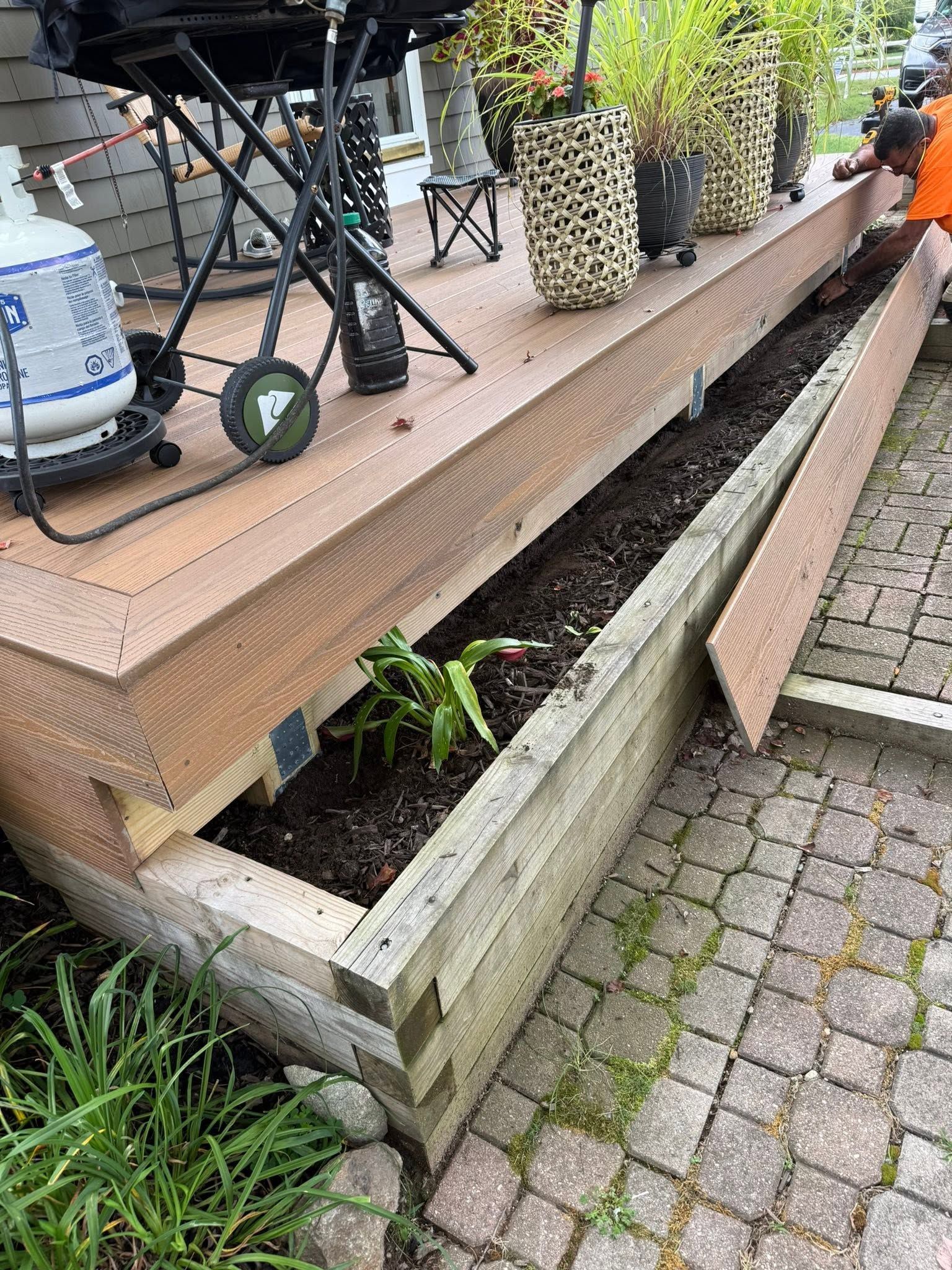 Raised planter bed with dark soil and a small green plant beside a cobblestone path and wooden deck edge