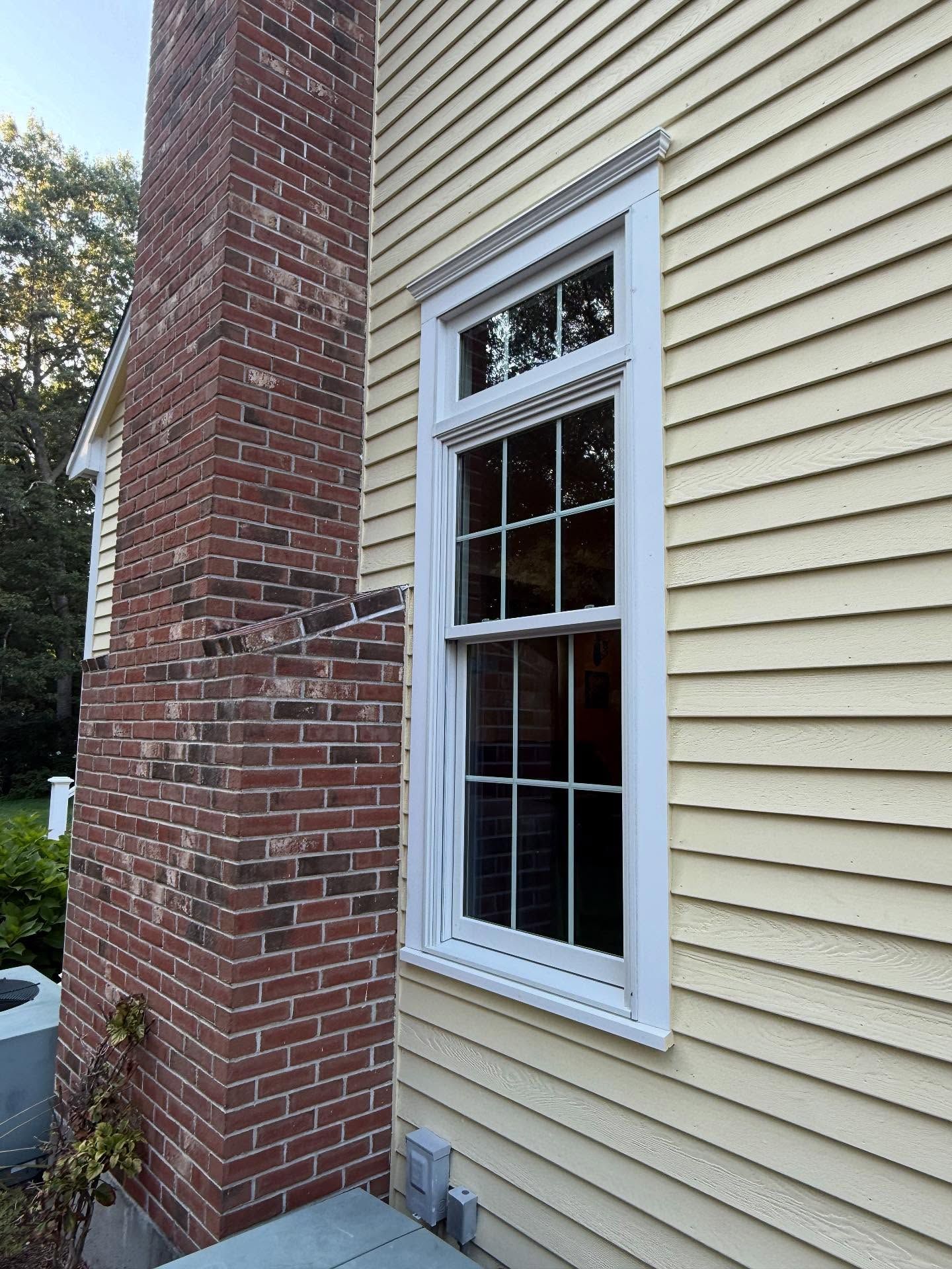 Brick chimney beside a beige-sided house with a white-trimmed window and visible exterior venting