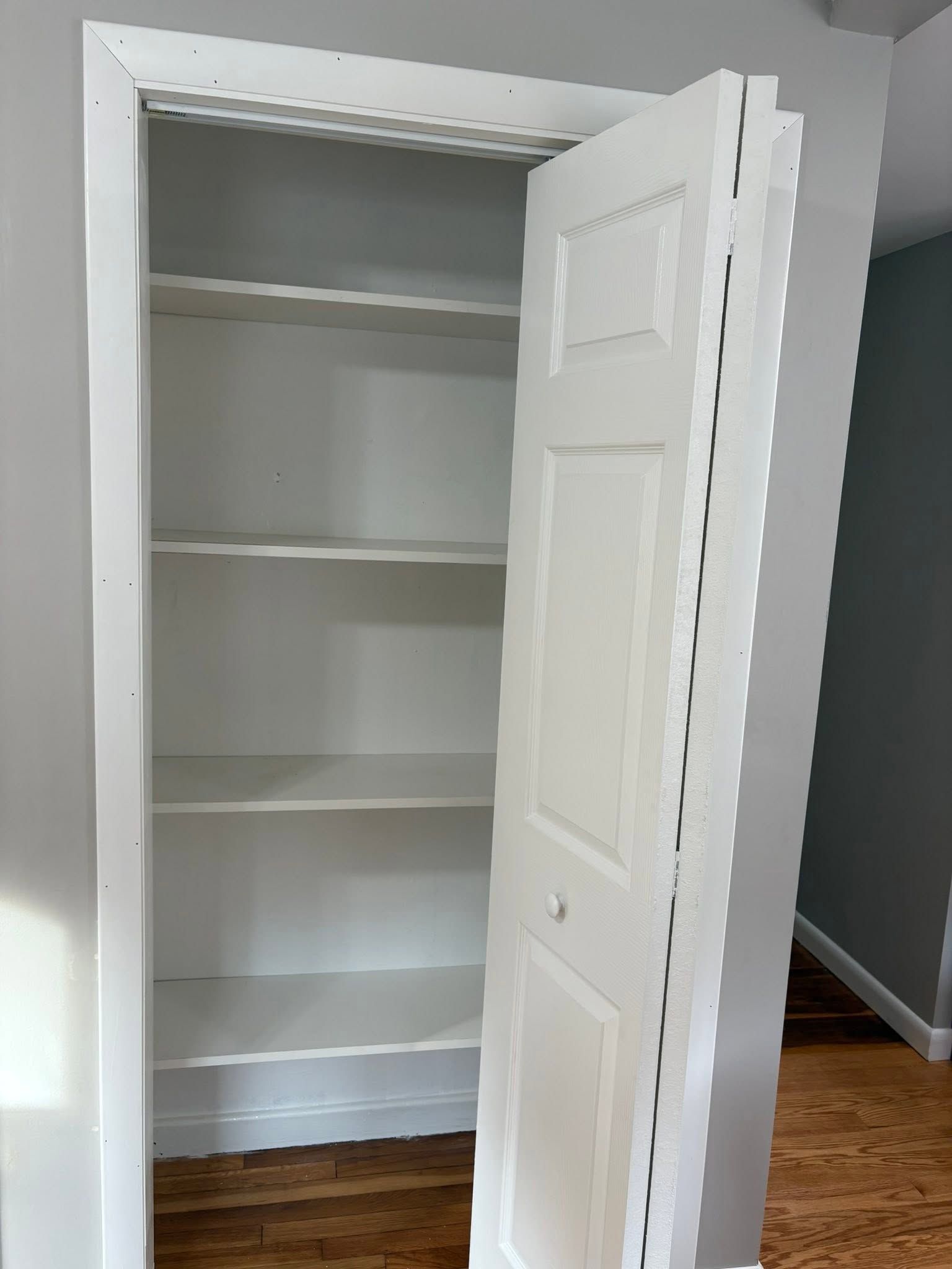 Open white closet with shelves and a partially open door in a room with hardwood floor
