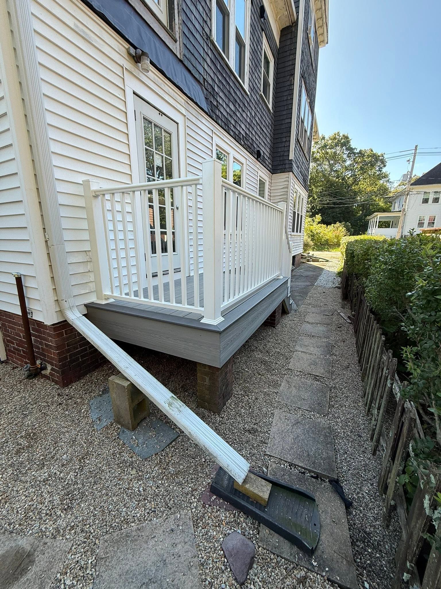 White porch with railing beside a steep gravel path and hedge next to a house.
