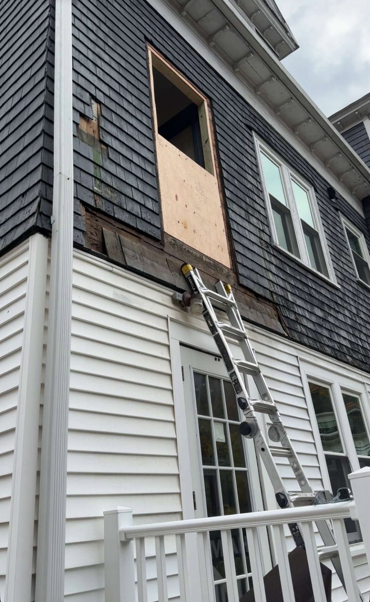 Ladder leaning to an open second-story window on a gray house with plywood covering the opening