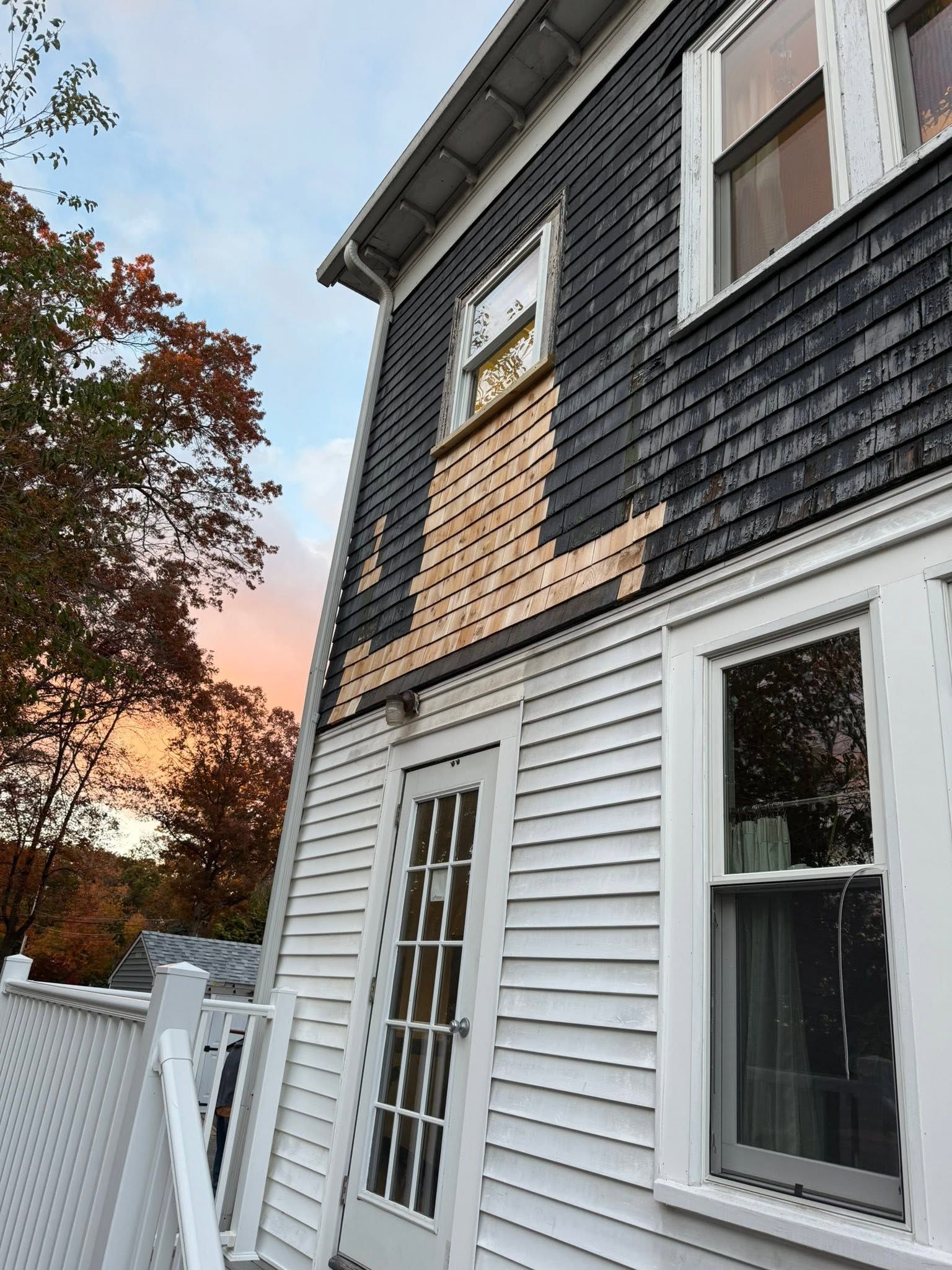 Two-story house with white siding and dark shingles at sunset, seen from the side with a fence and trees.