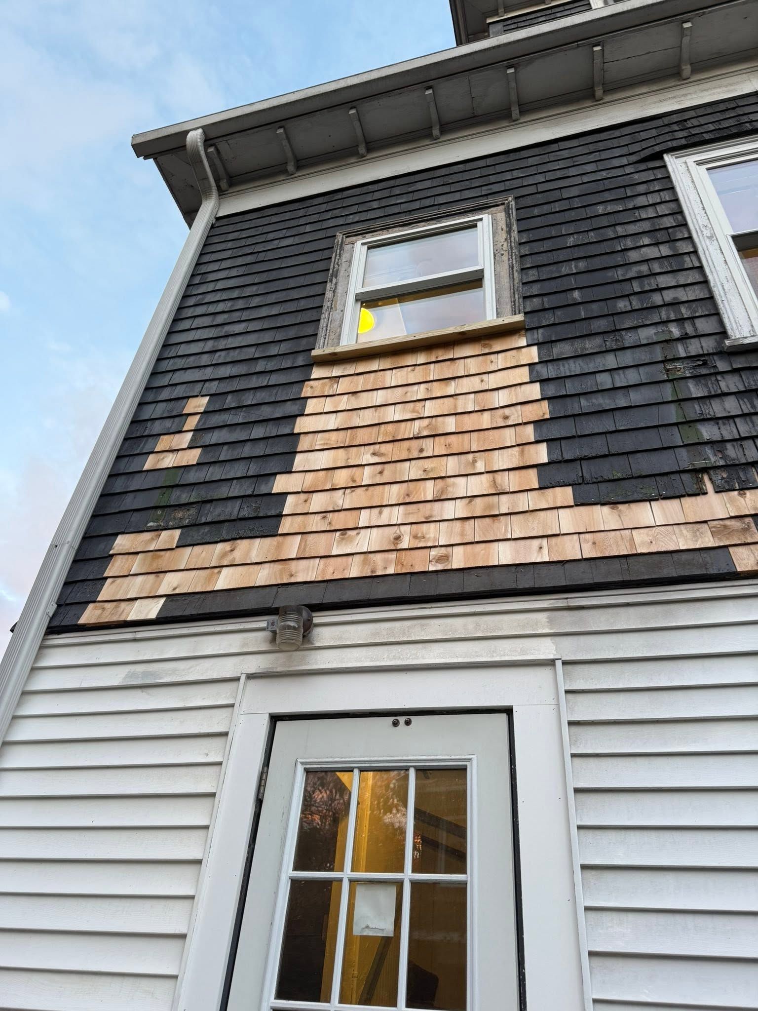 Exterior of a house with gray siding, black shingles, tan shingle patch, and two windows seen from below