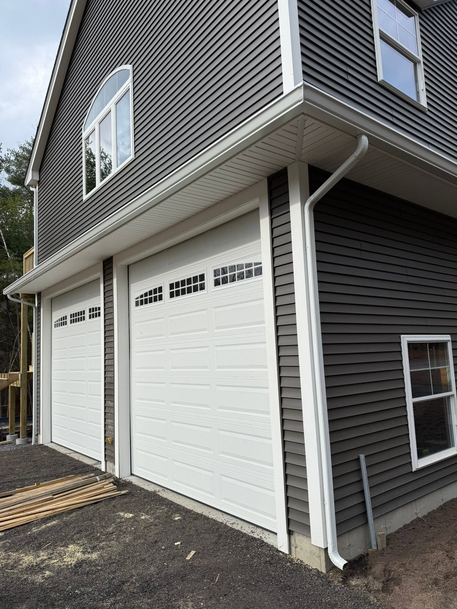 Two-story gray house with white garage doors and siding, viewed from the side outdoors