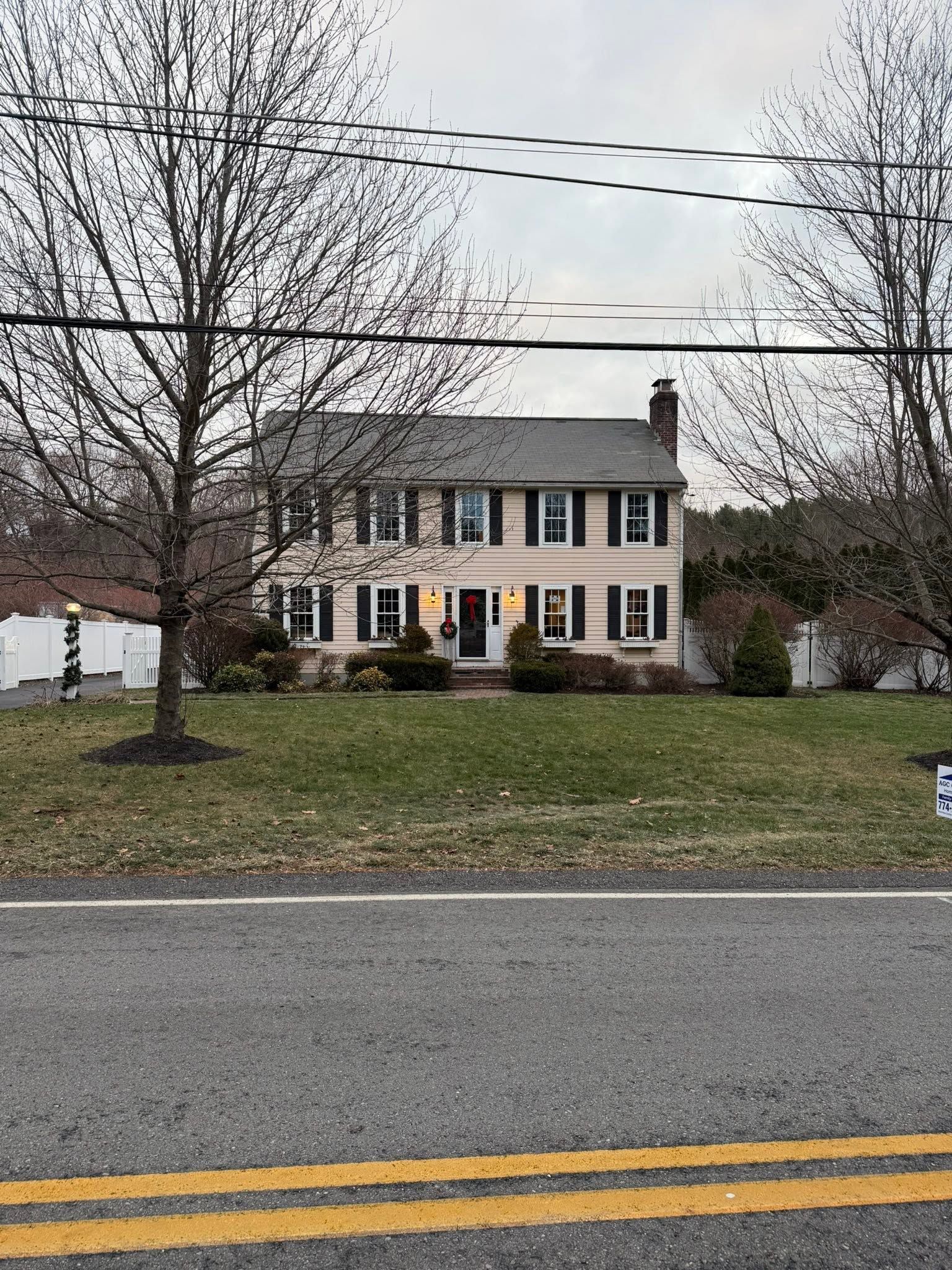 Two-story white house with black shutters and a front lawn beside a road on an overcast day