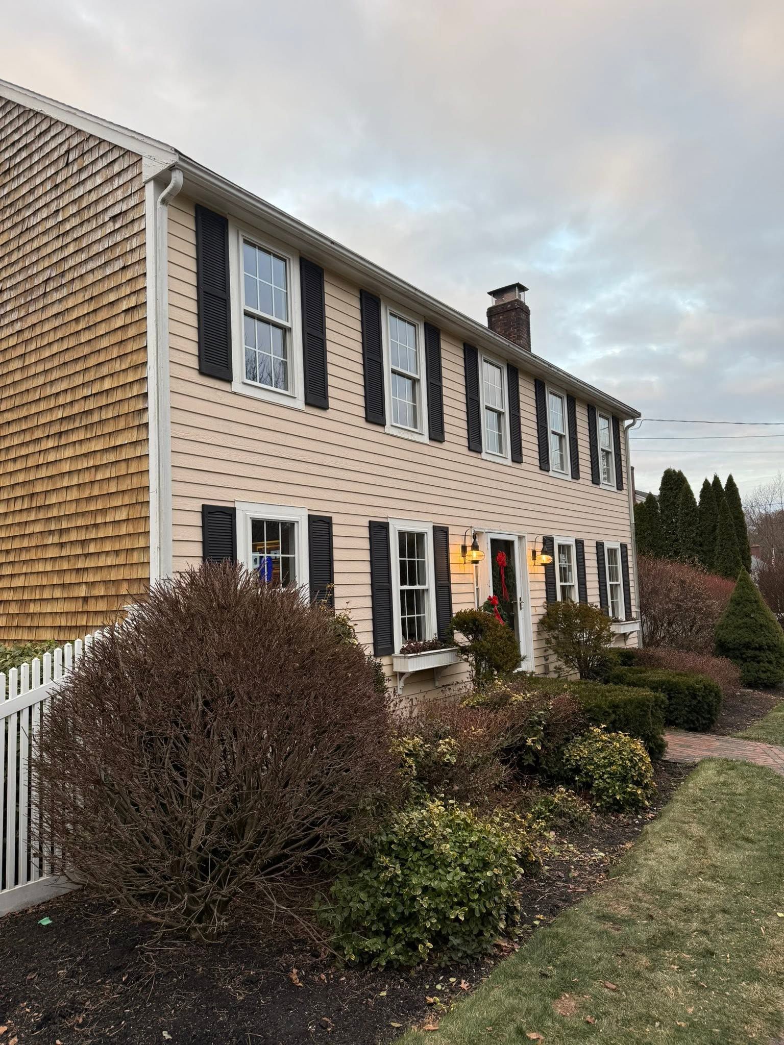Two-story tan house with black shutters, white trim, and a front garden under an overcast sky