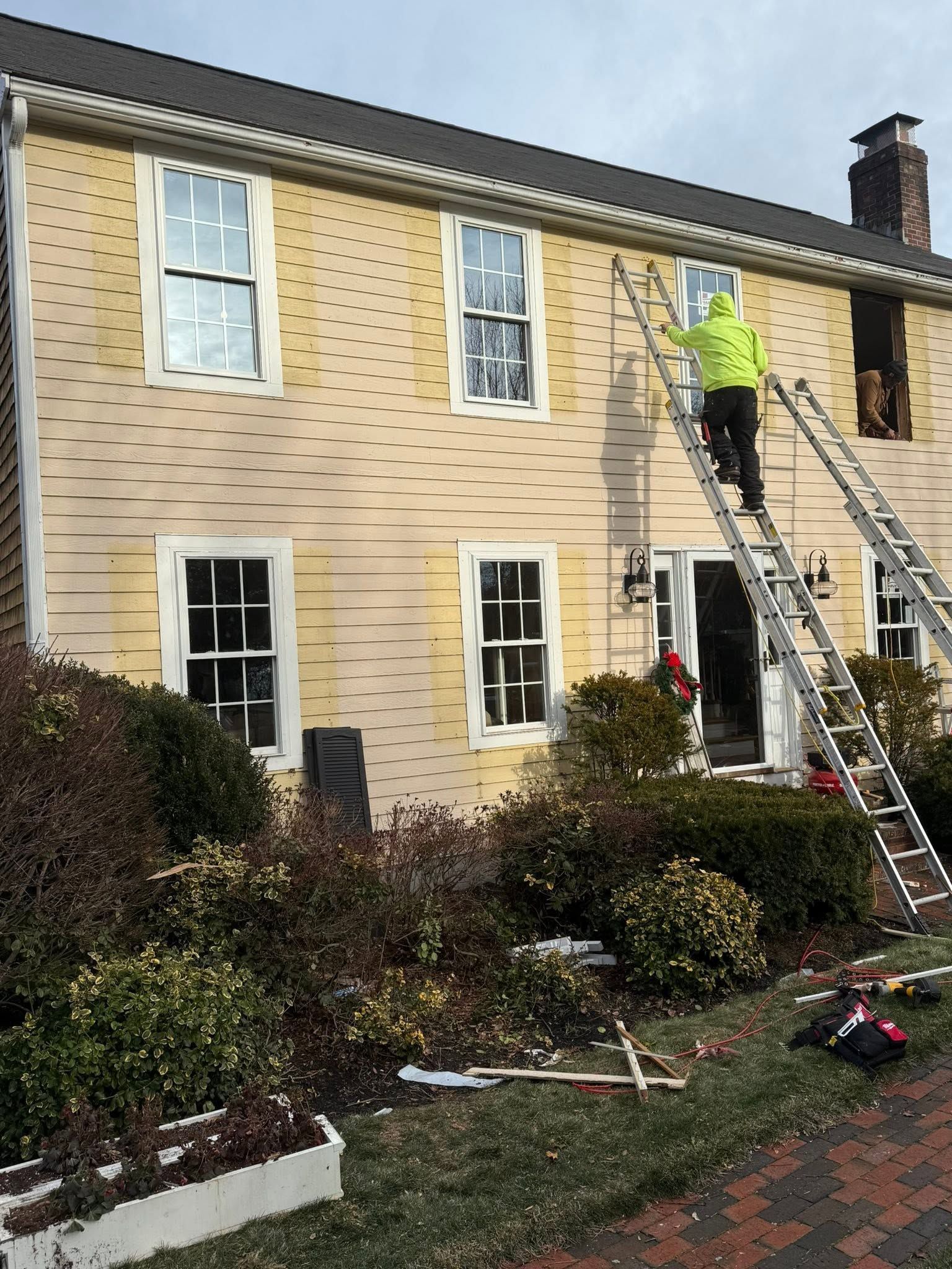 Worker on ladder painting the upper front of a cream house with white-trimmed windows and garden below