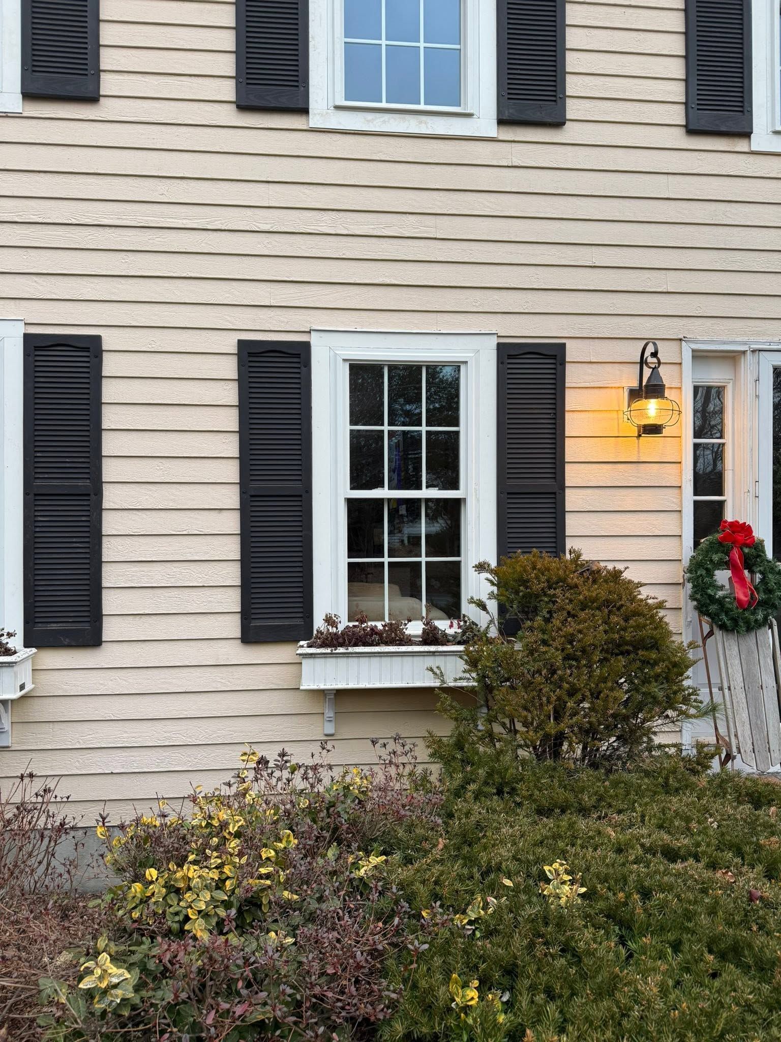 Suburban house exterior with beige siding, black shutters, a lit porch lamp, and flower boxes beneath the windows