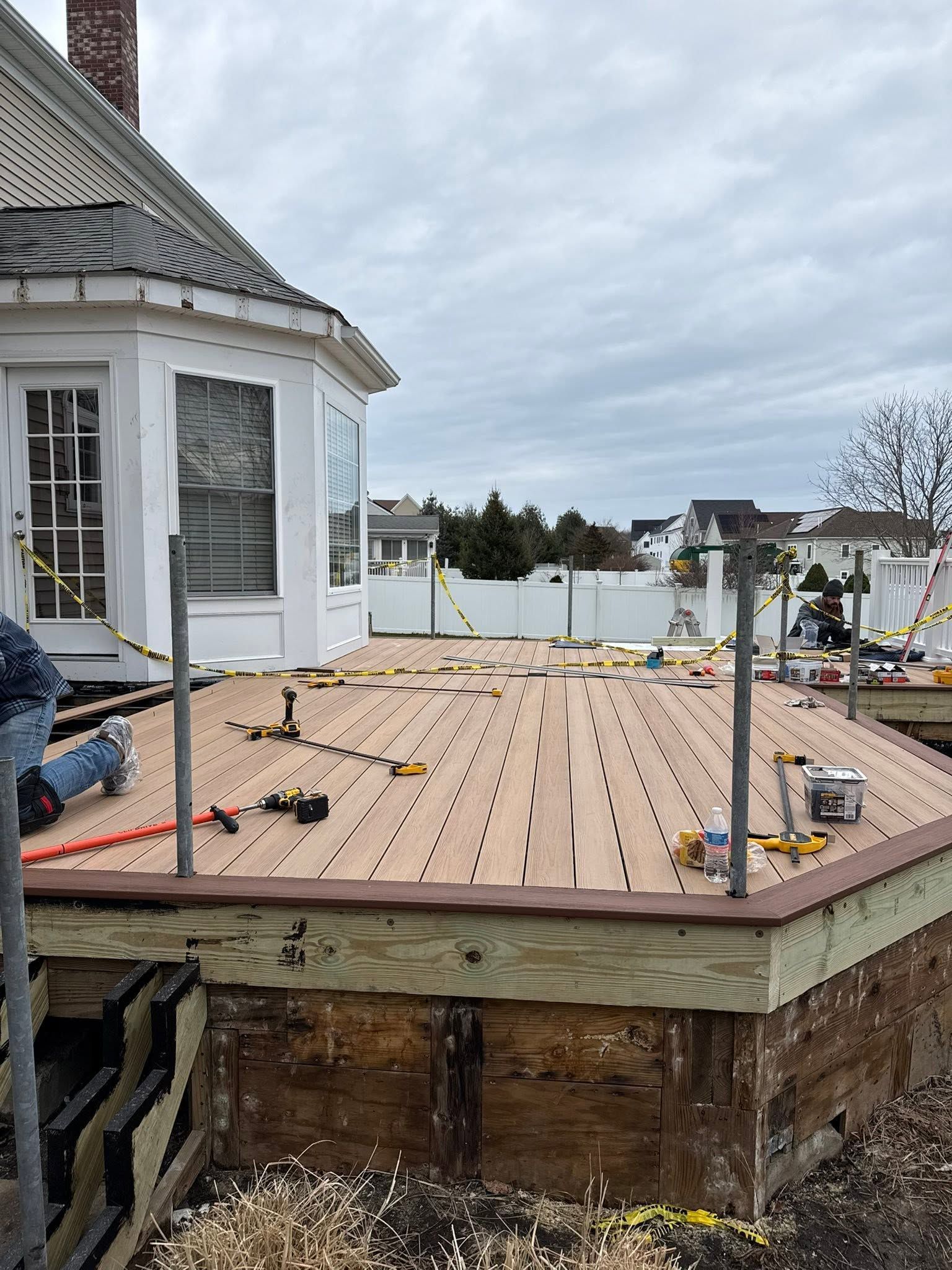 Deck under construction beside white house with exposed framing, tools, and cloudy sky