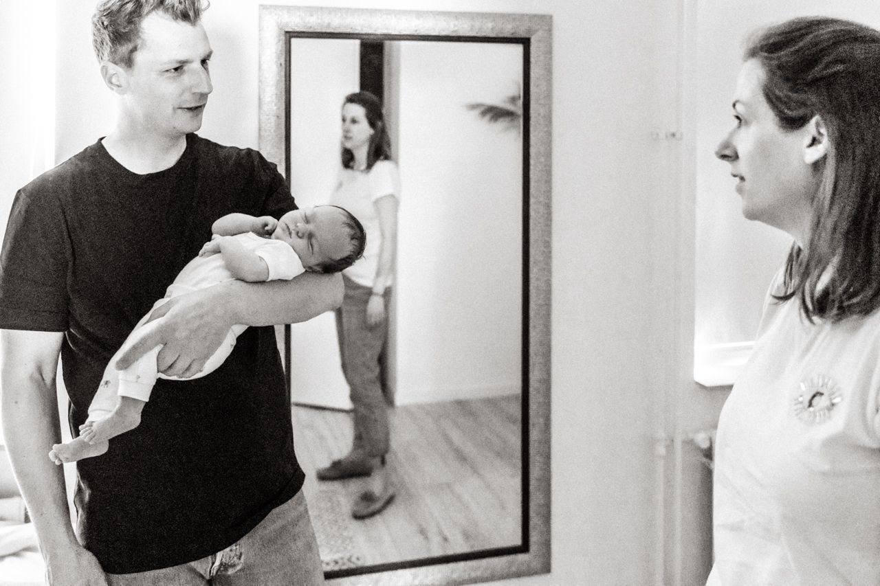 Een man houdt een baby voor de spiegel terwijl een vrouw toekijkt.