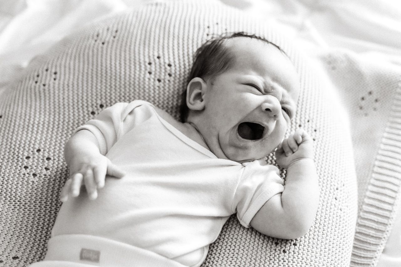 Een baby geeuwt terwijl hij op een deken op een bed ligt.