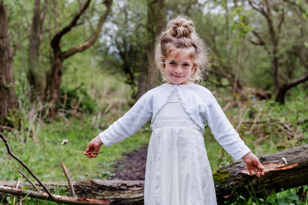 Een klein meisje in een witte jurk staat op een boomstam in het bos.