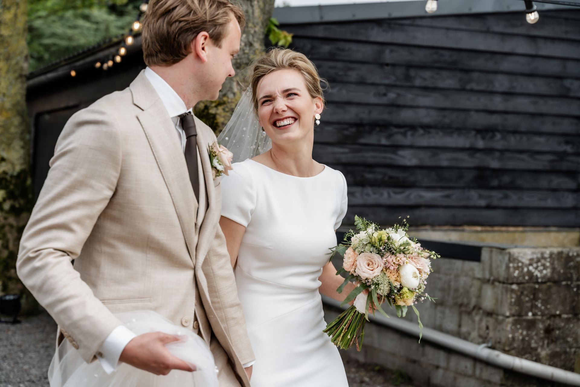 Een bruid en bruidegom houden elkaars hand vast en lachen voor een houten gebouw.