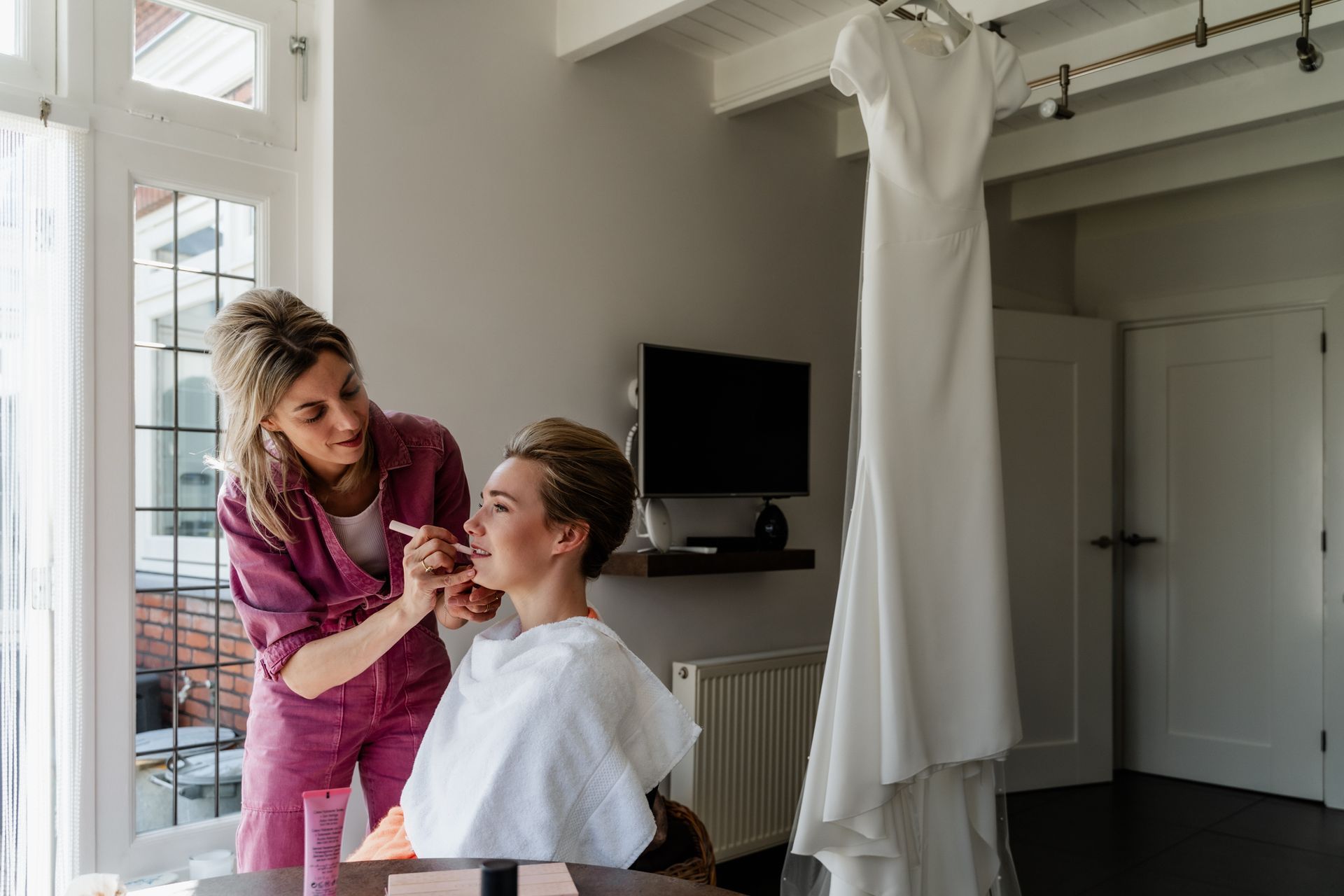 Een vrouw laat zich opmaken in een kamer waar een trouwjurk aan de muur hangt.