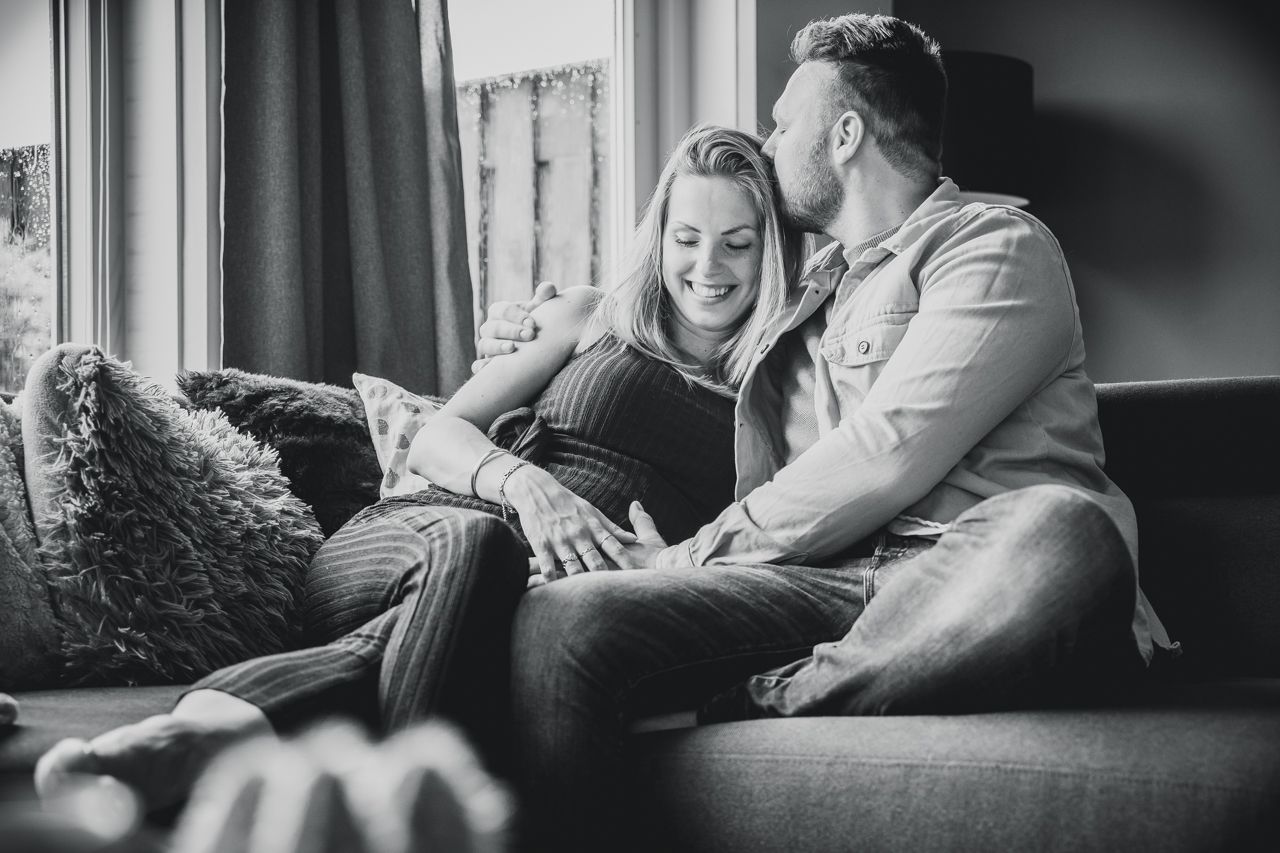 Een man kust een zwangere vrouw op het voorhoofd terwijl hij op een bank zit.