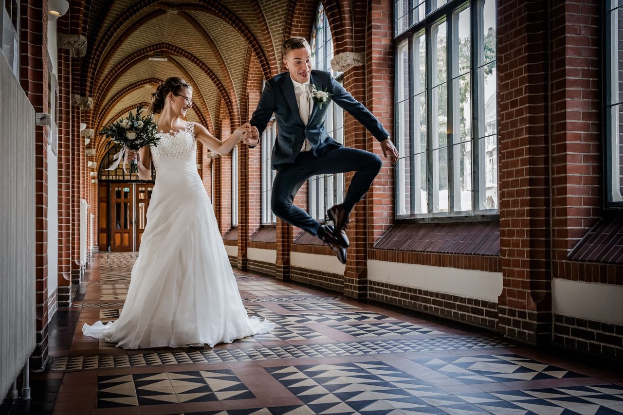 Een bruid en bruidegom lopen door een kloostergang en de bruidegom maakt een vreugdesprong.