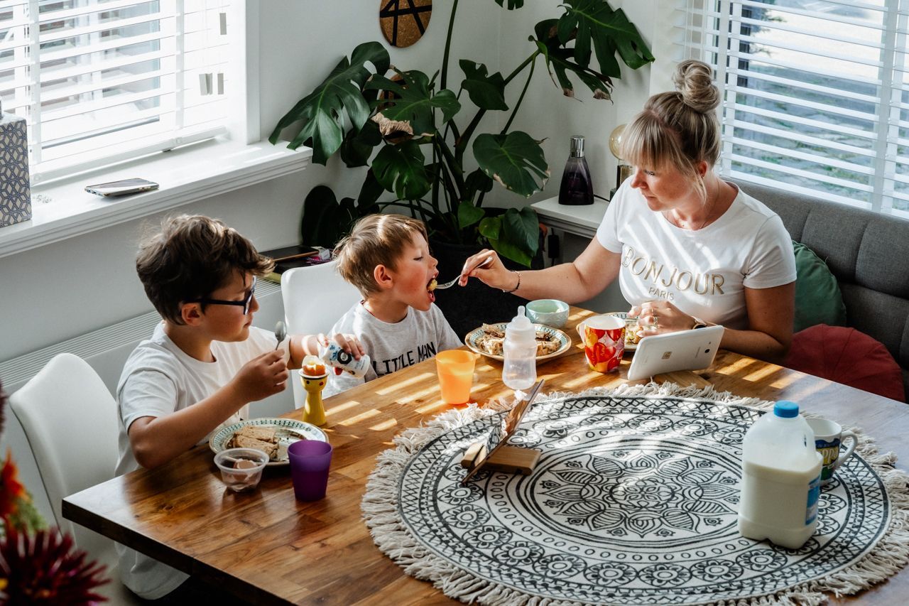 Een moeder met 2 jongetjes zitten aan de ontbijttafel en de moeder geeft 1 jongetje eten.