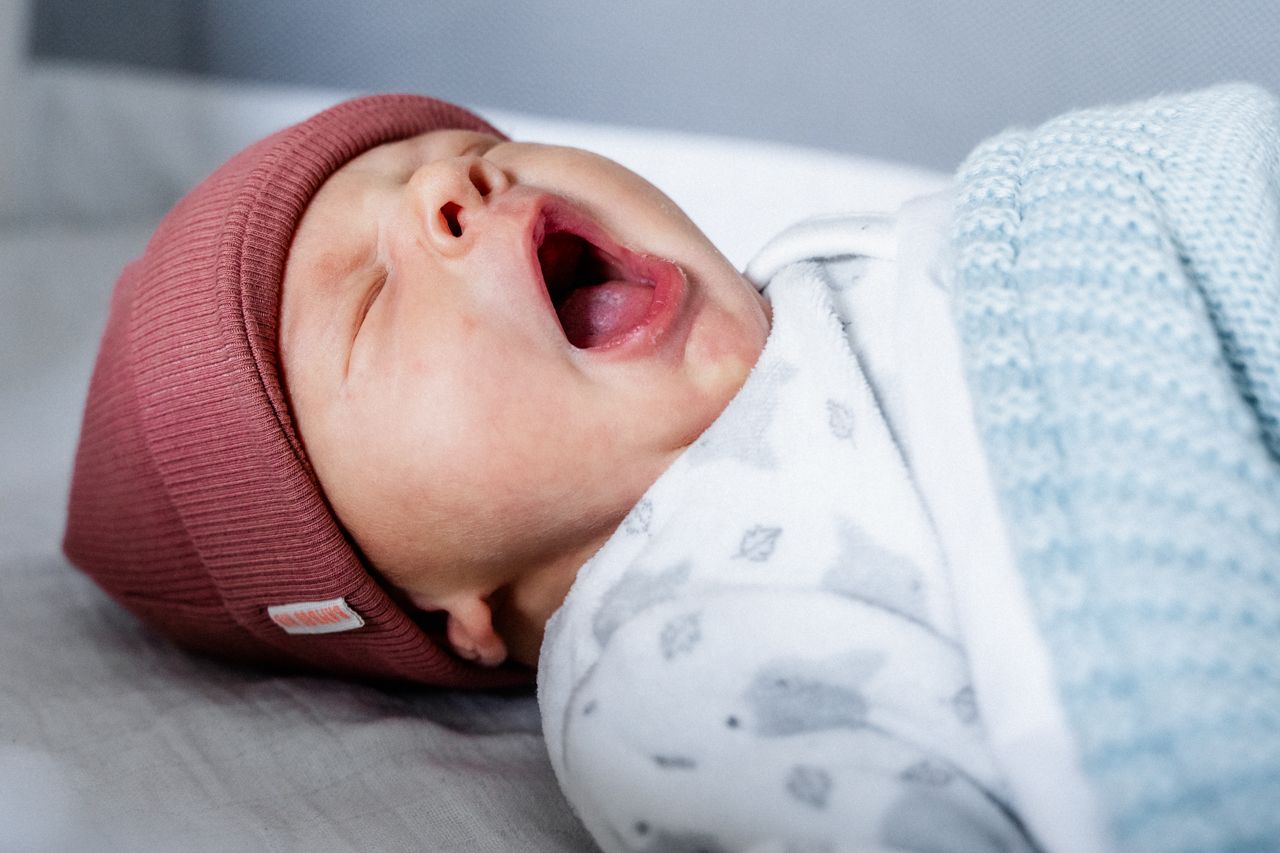 Een baby met een mutsje op geeuwt terwijl zij op bed ligt.