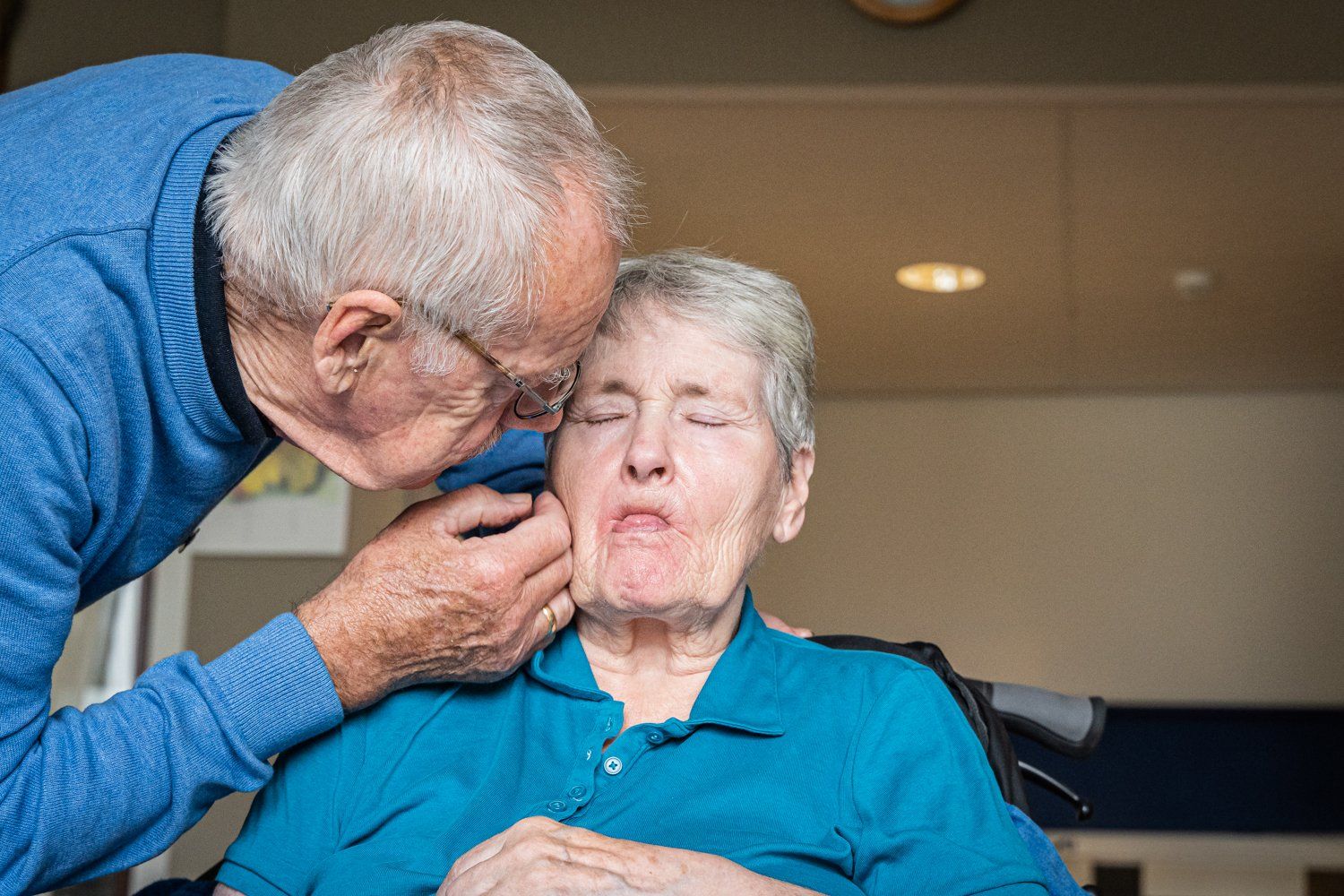 Een oudere vrouw in een rolstoel wordt door een man op de wang gekust.