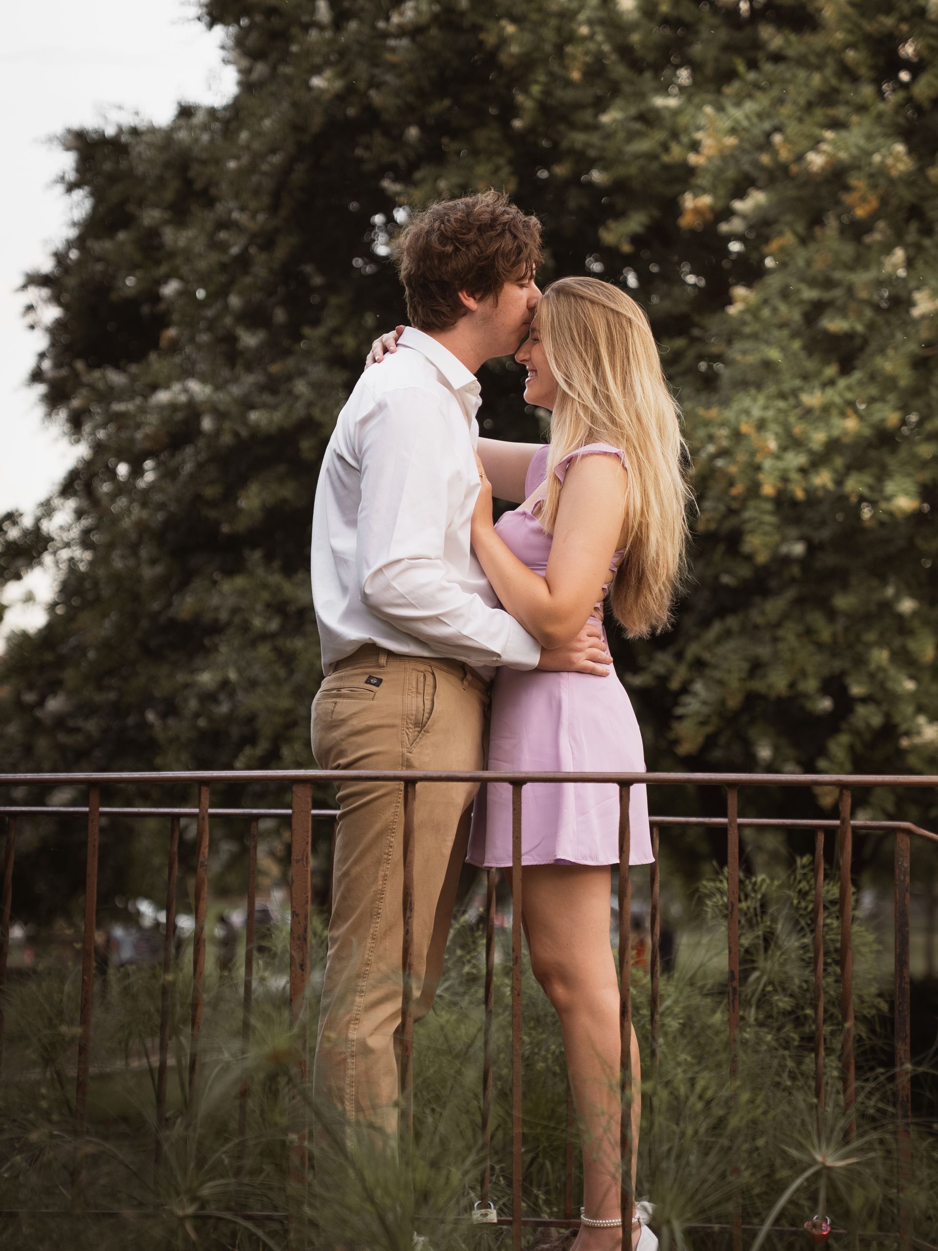 A man kisses his girlfriend on the forehead on a bridge in a park.