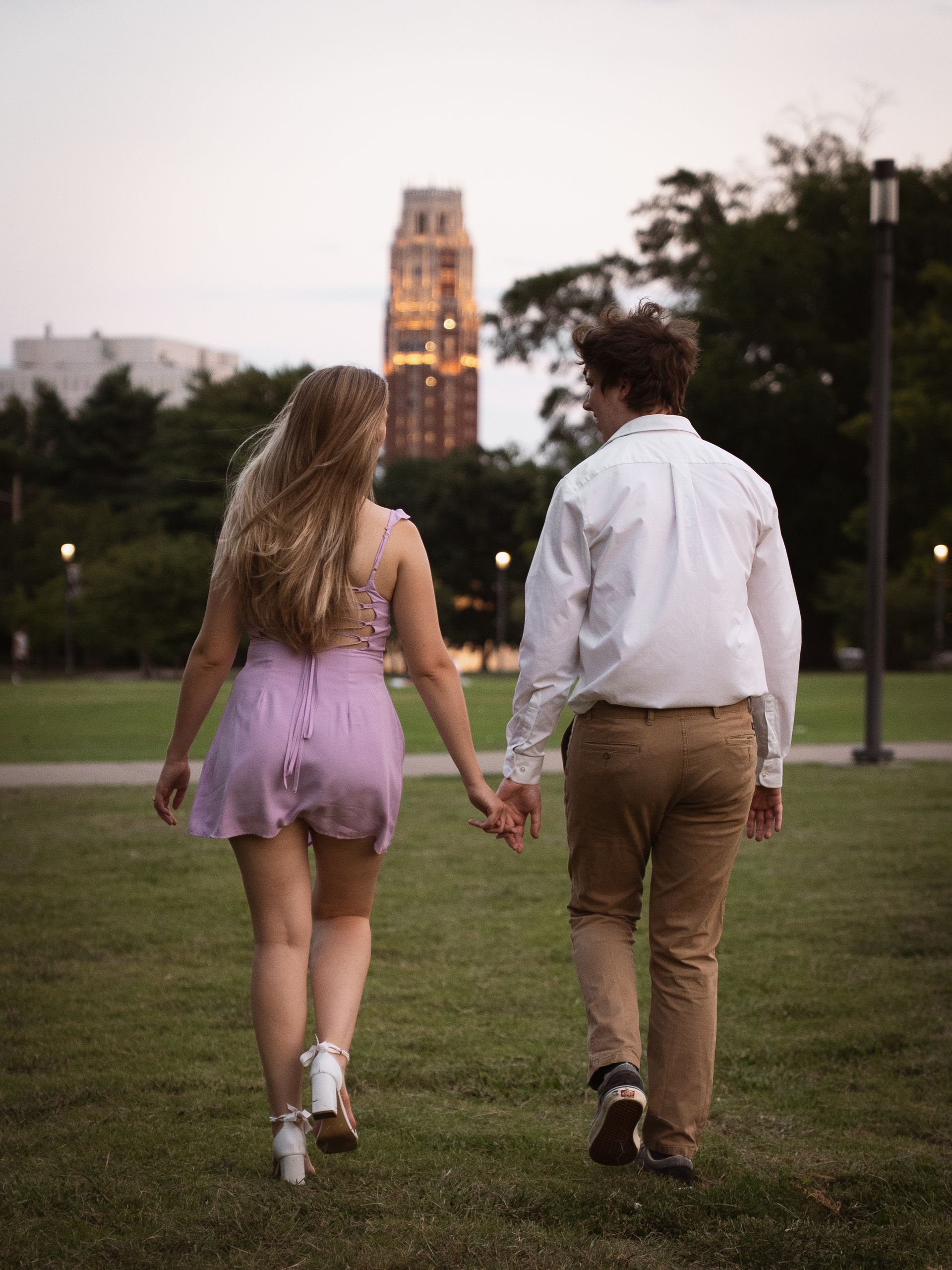 A recently engaged couple walks through a park, holding hands, with a beautiful building in the background.