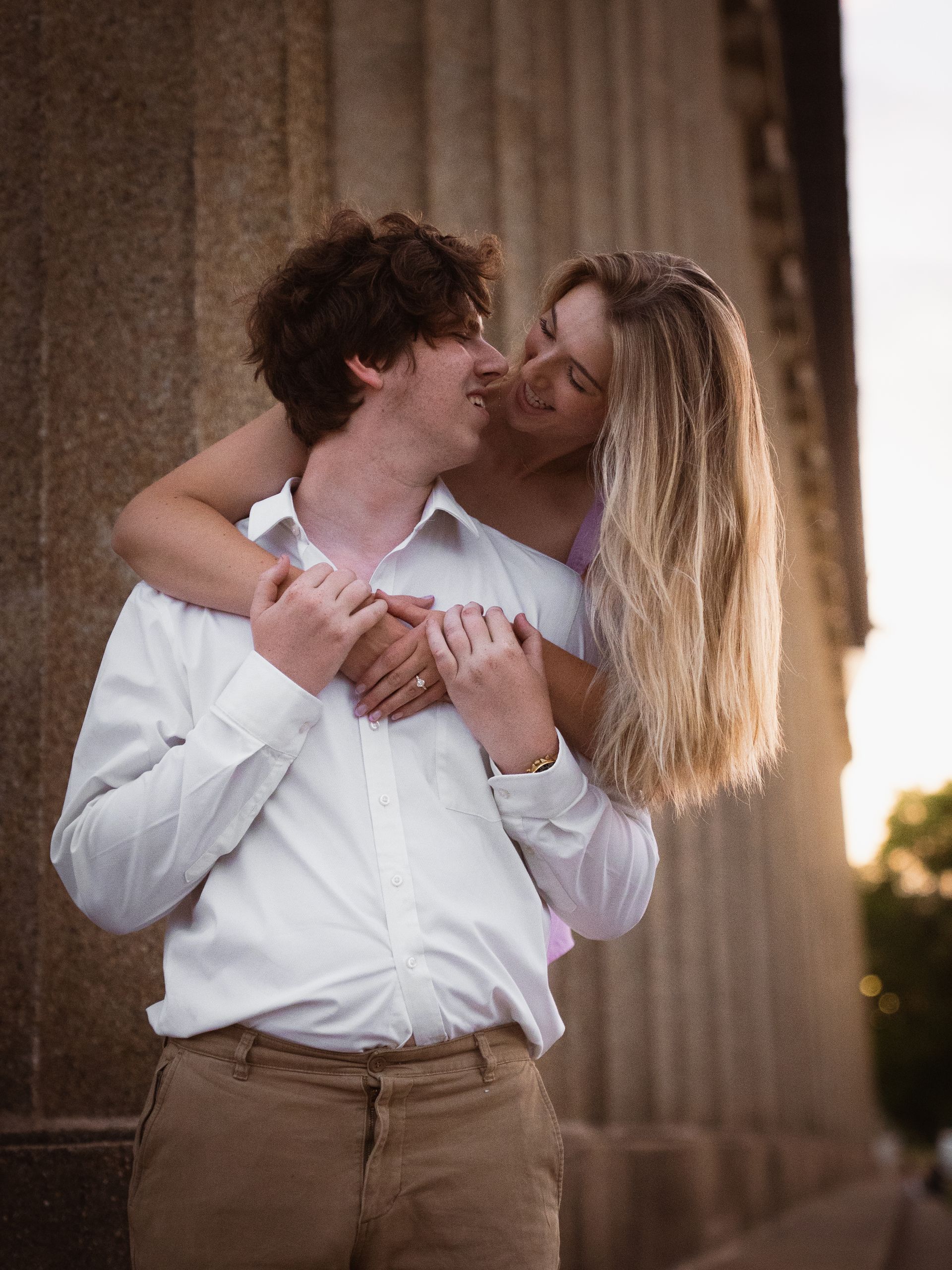 A woman leans over her fiancee's shoulder, smiling at him at sunset.