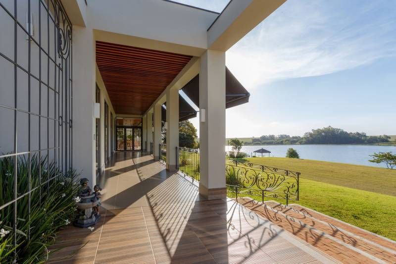 Varanda longa com vista para o lago; casa com ladrilhos marrons e cerca de ferro preta, dia ensolarado.