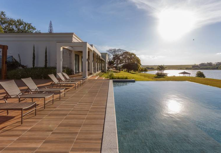 Espreguiçadeiras ao lado de uma piscina infinita com vista para um lago, com um edifício branco moderno sob um céu ensolarado.
