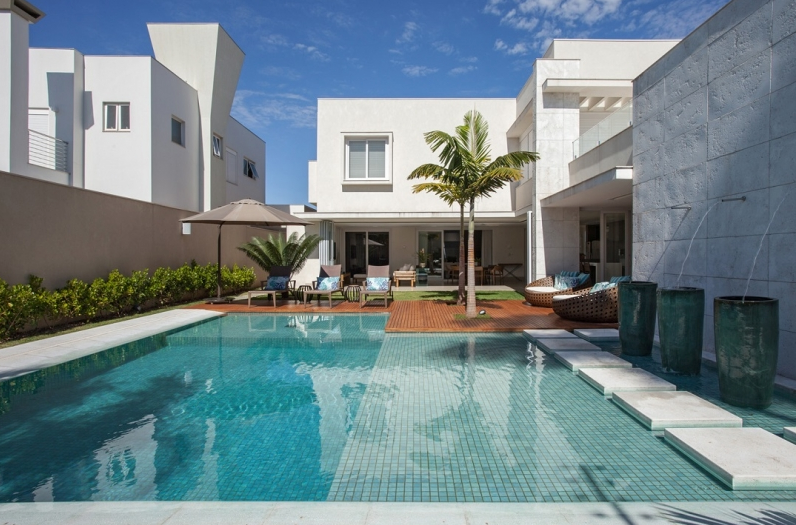Casa branca moderna com piscina, espreguiçadeiras e uma pequena palmeira em um pátio ensolarado.