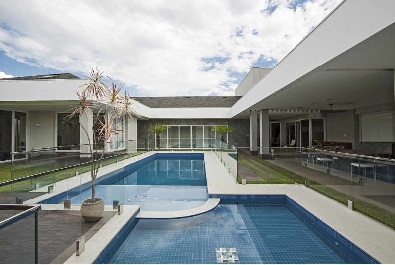Casa moderna com piscina retangular, água azul, grades de vidro e assentos ao ar livre.