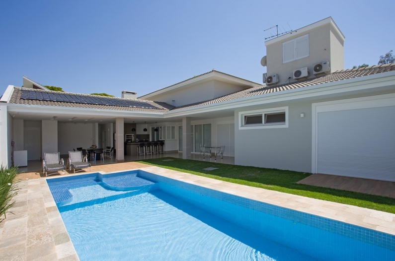 Quintal com piscina, pátio, churrasqueira e casa cinza claro em um dia ensolarado.