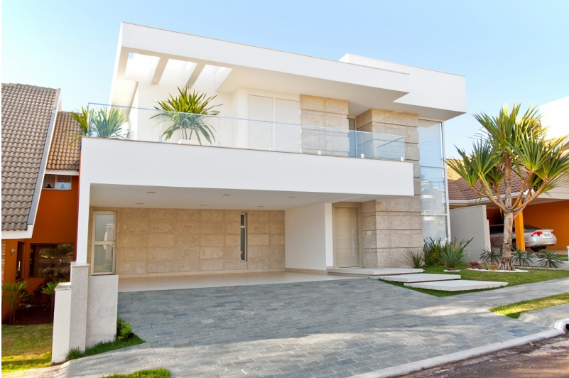 Casa moderna, branca, de dois andares, com fachada de pedra, varandas de vidro e entrada para carros. Palmeira à direita.