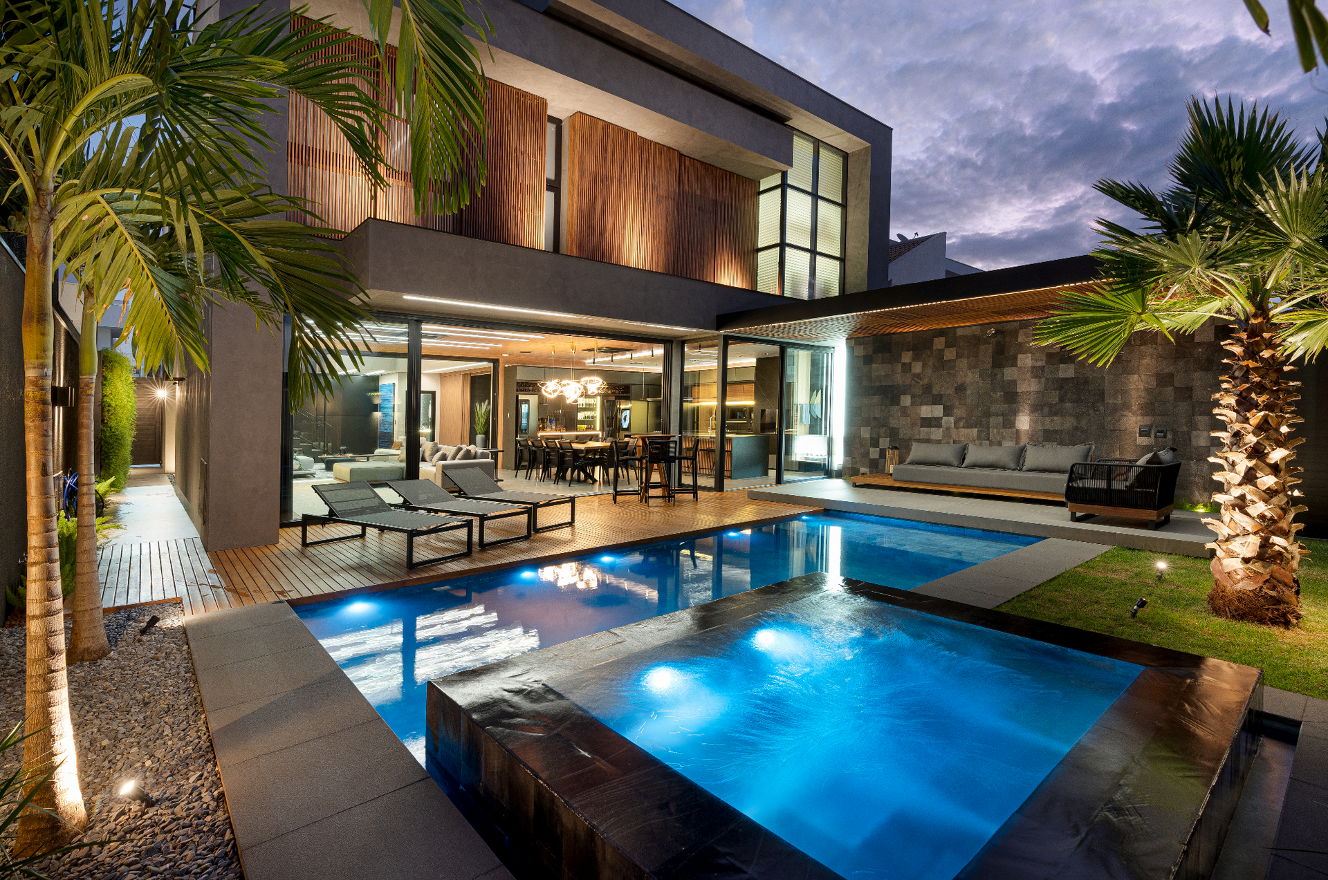 Casa moderna com piscina ao entardecer; água iluminada, palmeiras, deck, área de estar.