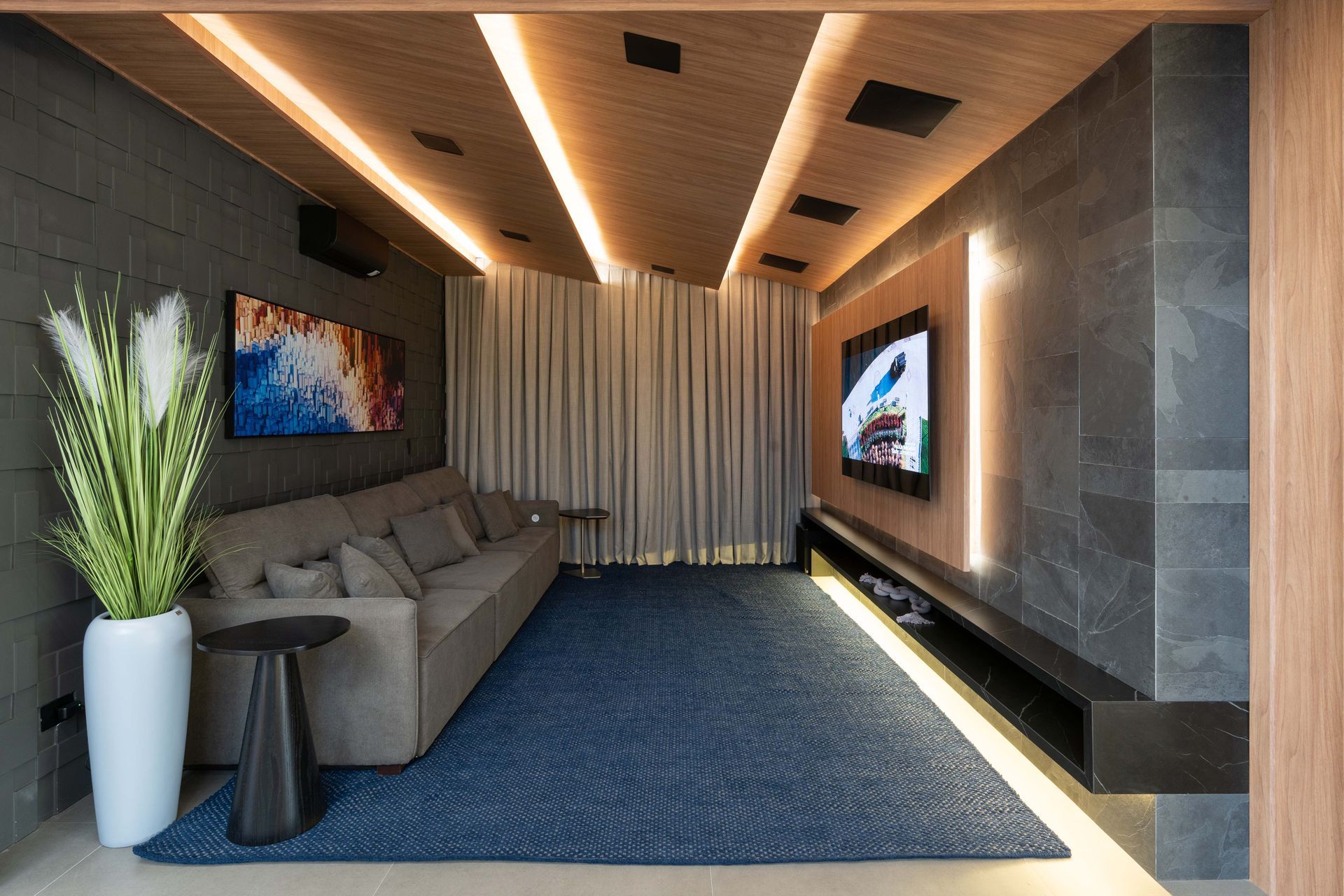 Home theater aconchegante com sofá cinza, tela grande, tapete azul e iluminação decorativa.