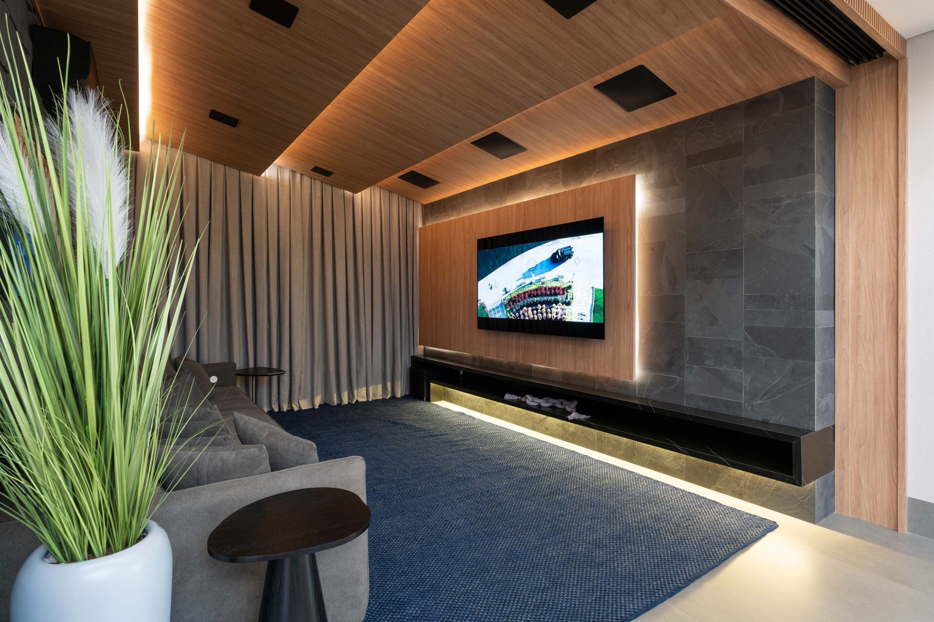 Home theater aconchegante com uma TV grande, parede de pedra, teto de madeira e carpete azul. Planta alta em primeiro plano.