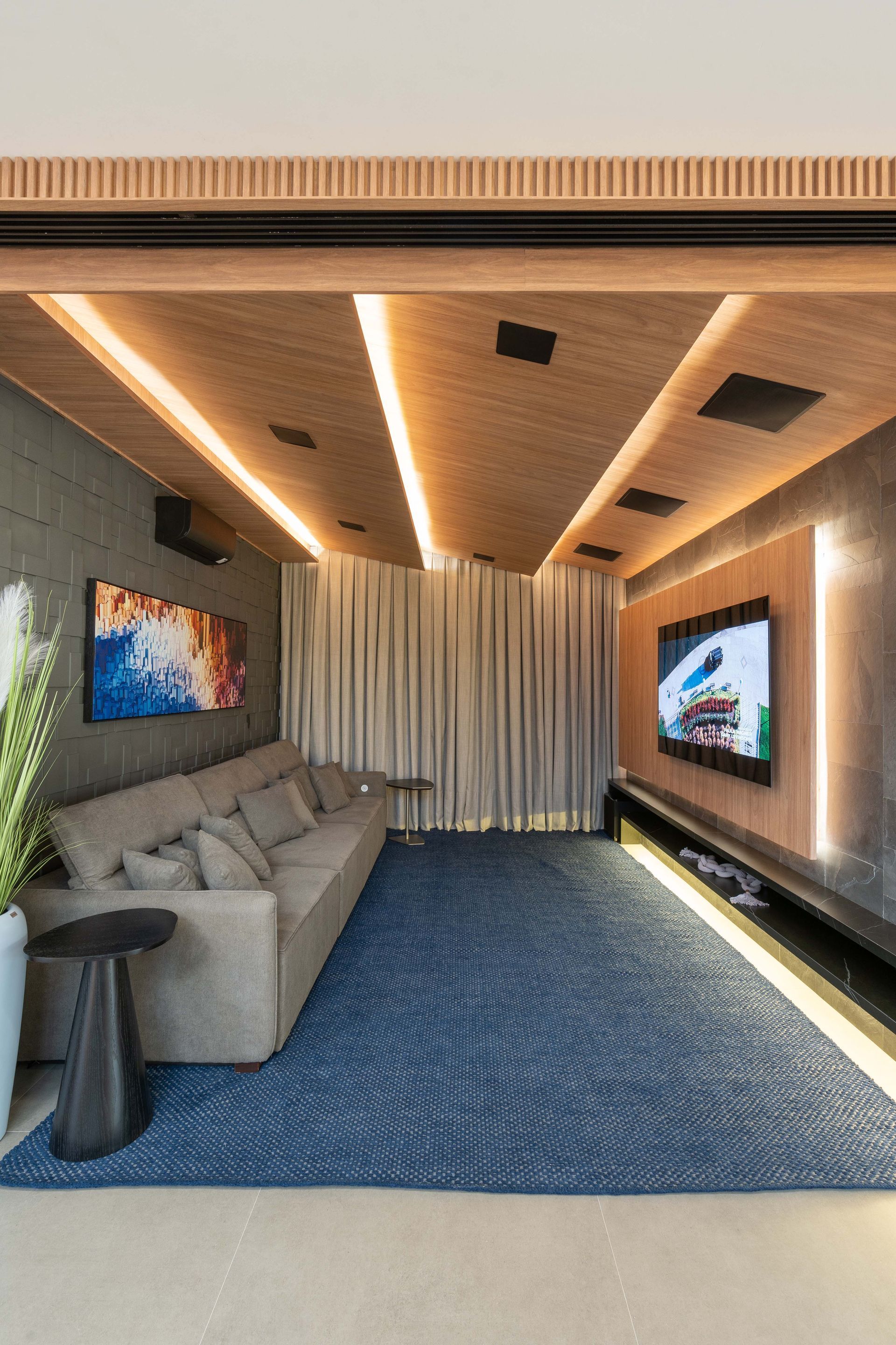 Home theater aconchegante: sofá cinza, tapete azul estampado, teto de madeira com luzes, TV grande e cortinas neutras.