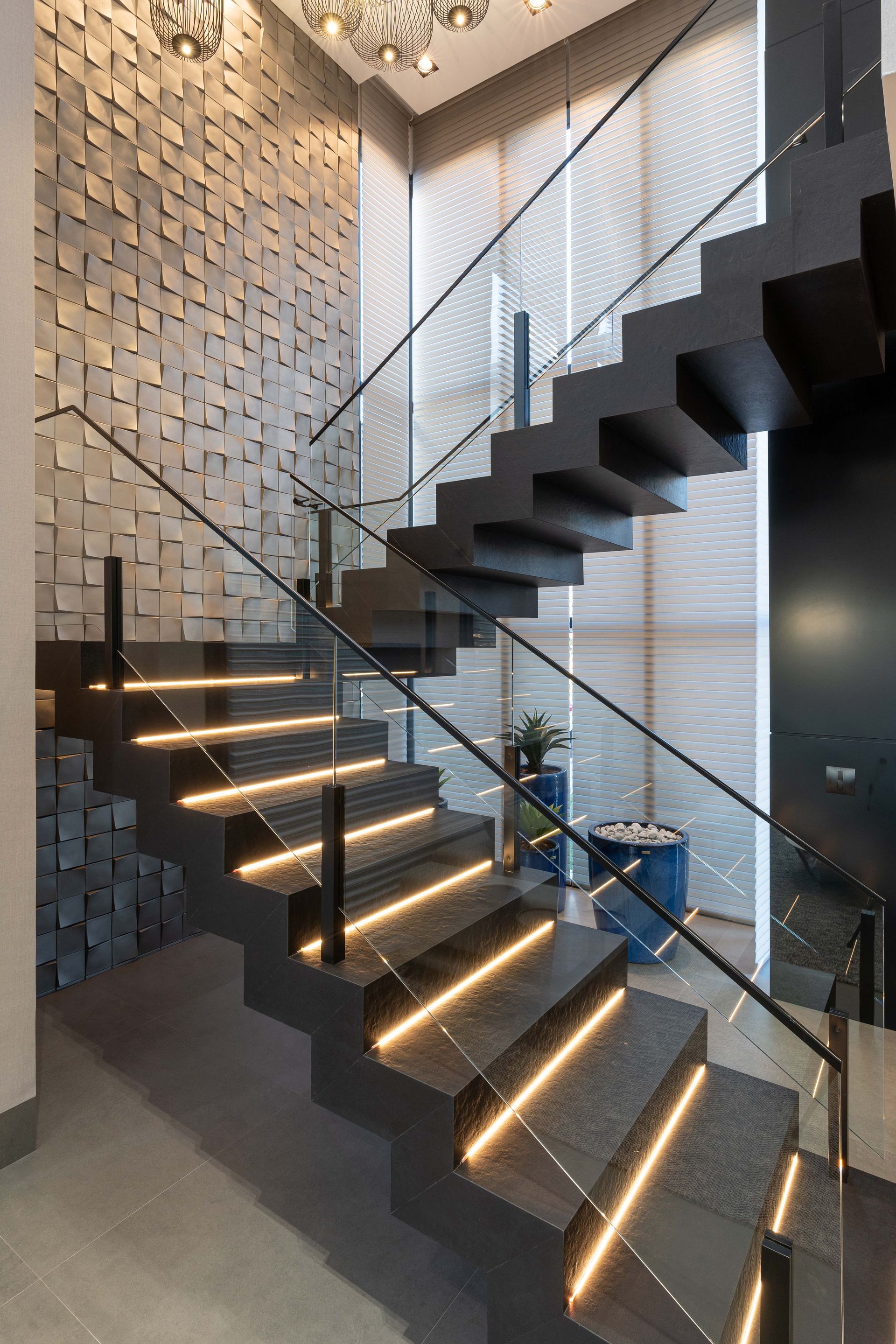 Escada moderna com degraus pretos, corrimão de vidro e iluminação integrada. Parede texturizada em pedra.