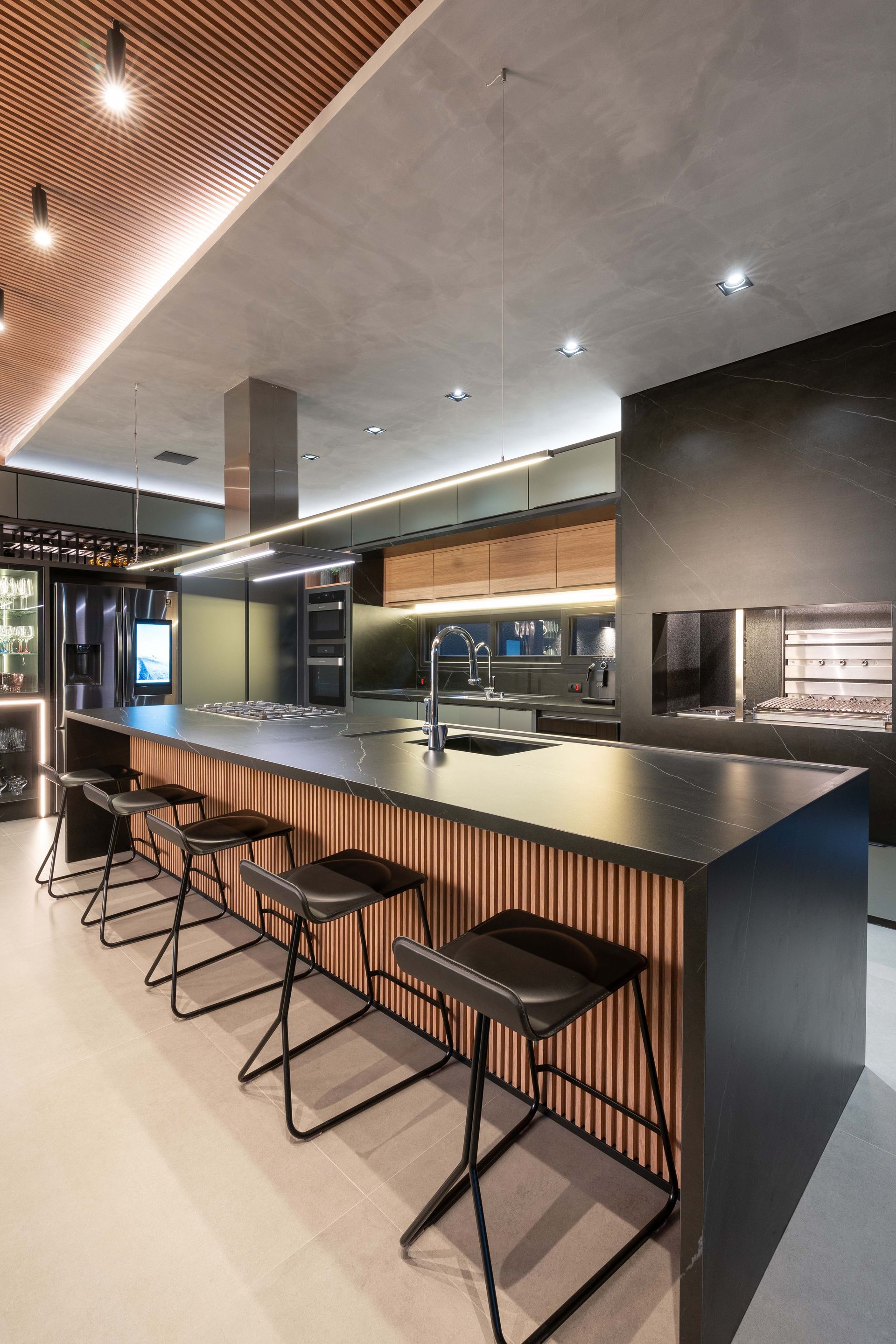 Cozinha moderna com bancadas pretas, detalhes em madeira, bancos de bar e churrasqueira embutida.