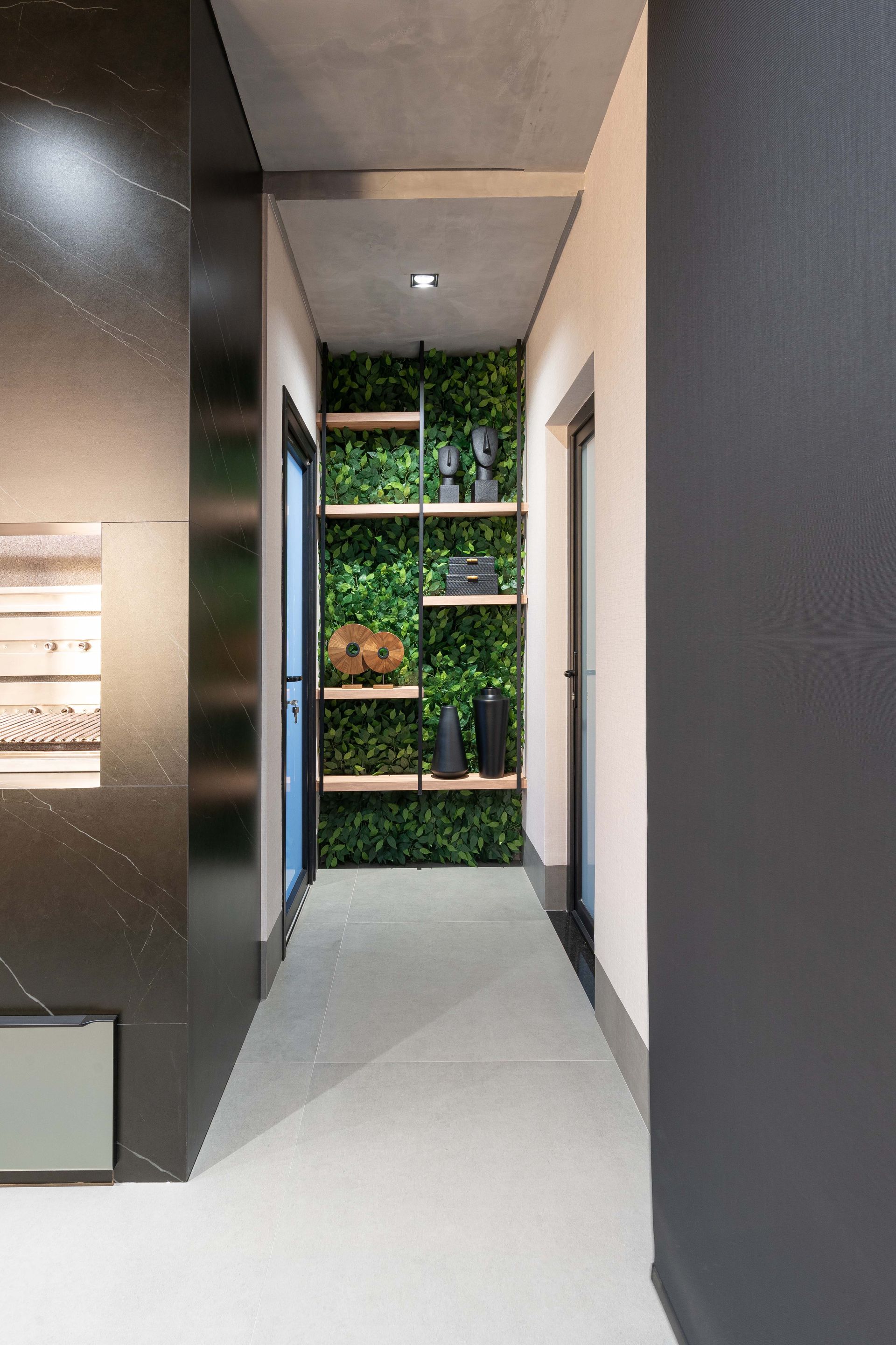 Corredor estreito com uma parede de plantas verdes, prateleiras de madeira com vasos pretos e uma porta escura.