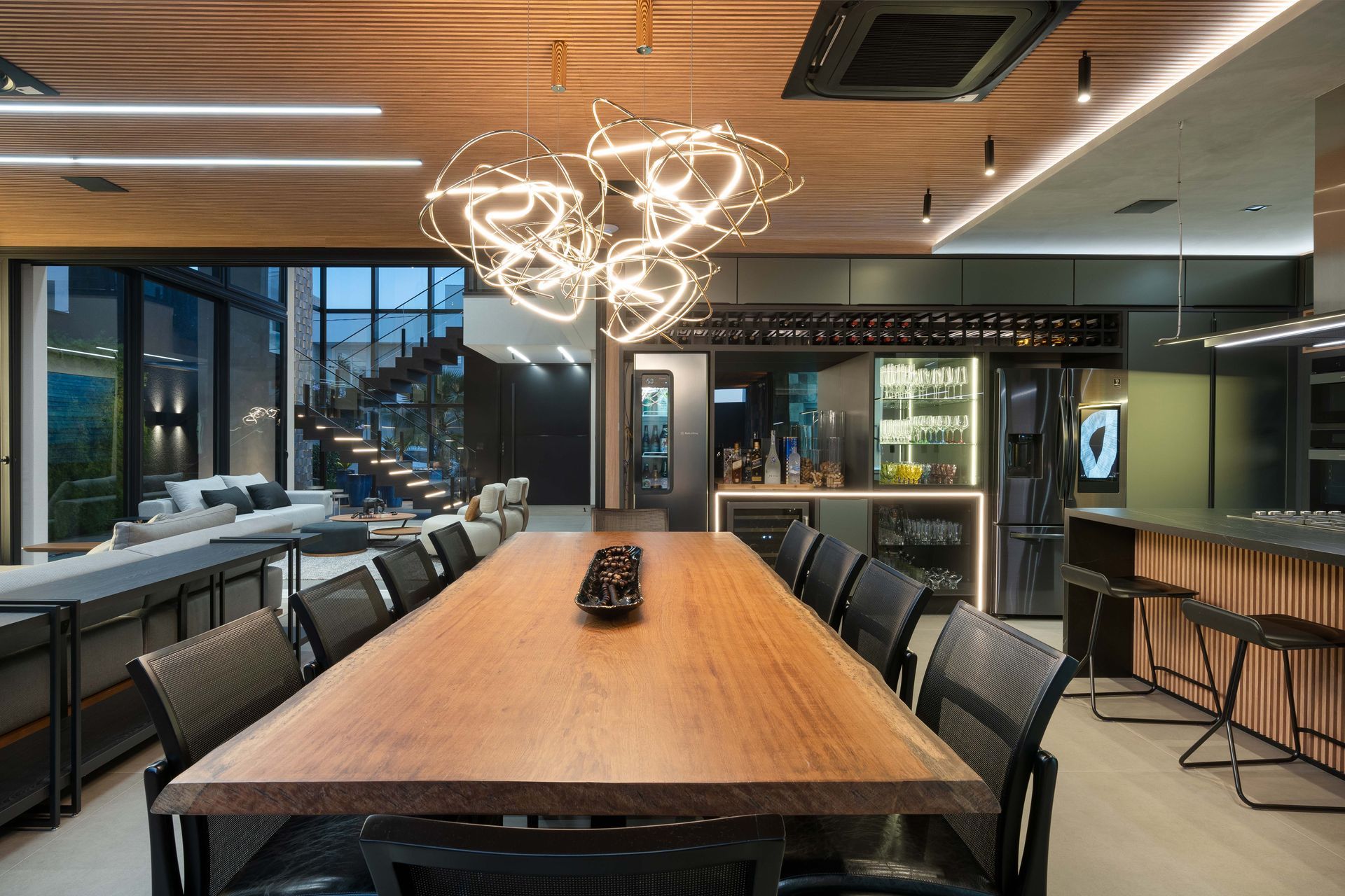 Uma sala de jantar moderna com uma longa mesa de madeira, cadeiras e um bar, com vista para uma escada.