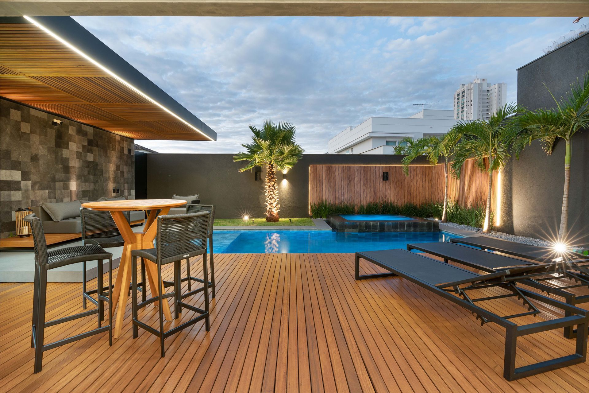 Área de piscina ao ar livre com deck de madeira, bar, espreguiçadeiras, piscina e plantas ao entardecer.