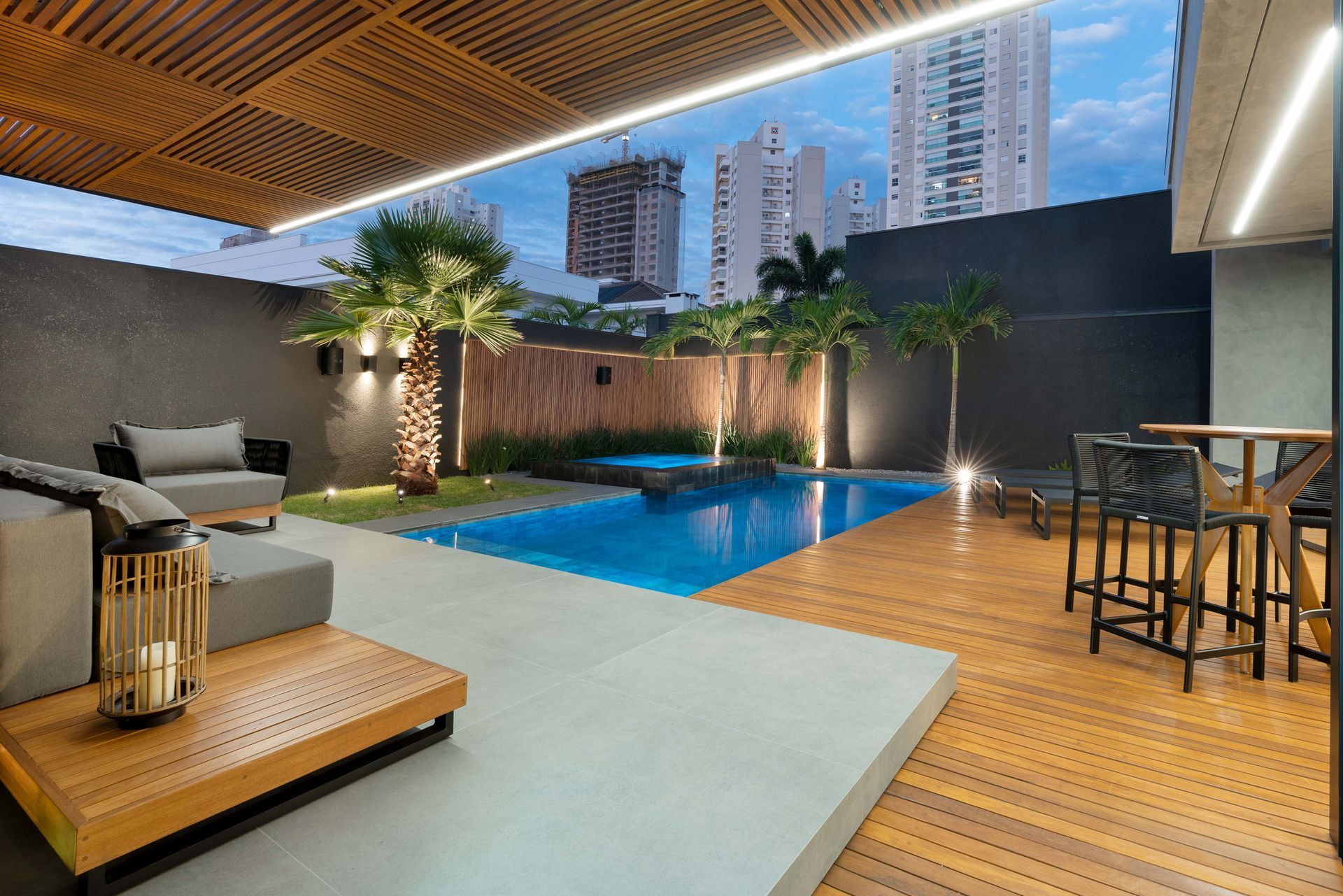 Área de estar ao ar livre com piscina, assentos e deck de madeira sob um teto de ripas com vista para o horizonte da cidade.